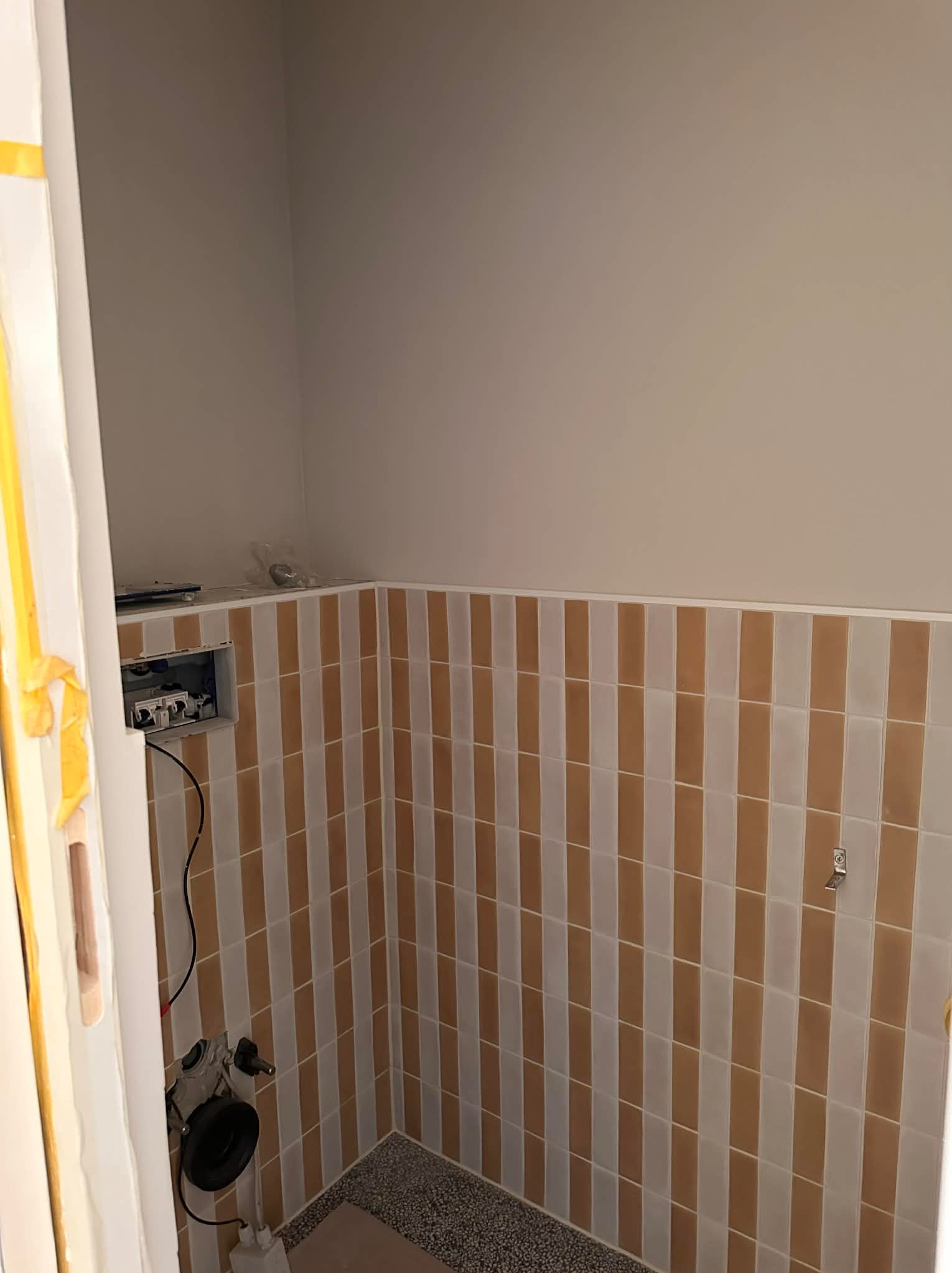 Wnętrze łazienki w trakcie remontu: ściany wyłożone pionowymi, brązowo-białymi płytkami, widoczna instalacja elektryczna i zabezpieczone futryny drzwiowe.