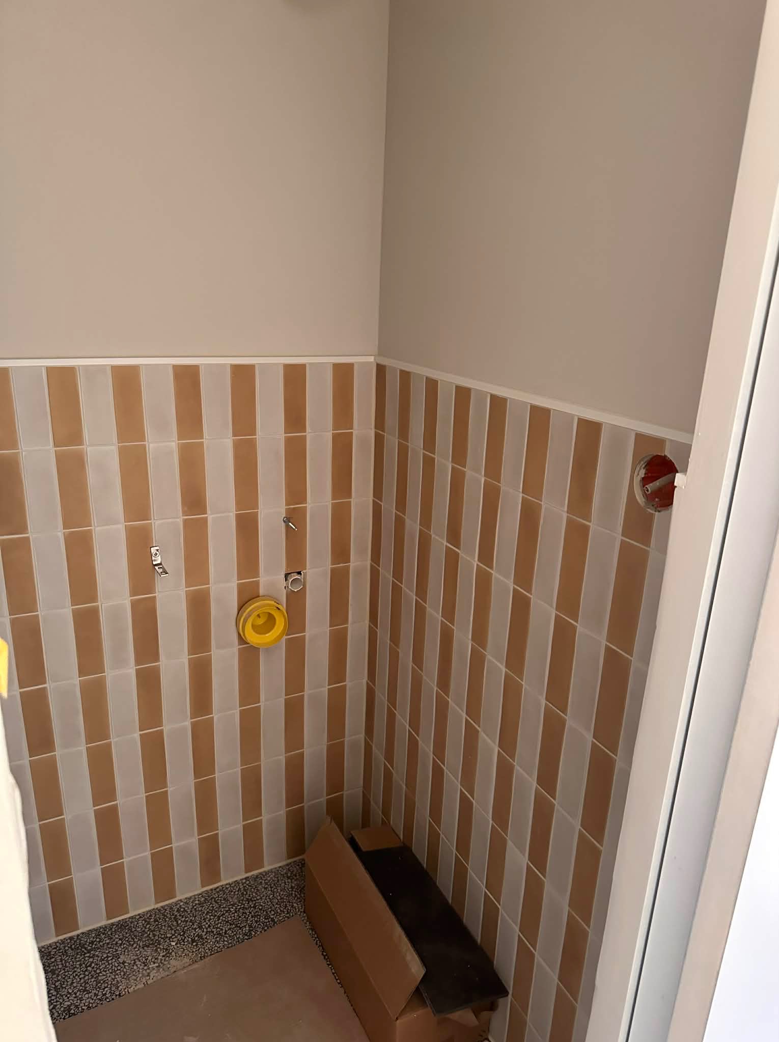 Ściana łazienki wyłożona pionowymi, brązowo-białymi płytkami. Widoczne przygotowane punkty instalacyjne i fragment podłogi przed wykończeniem.