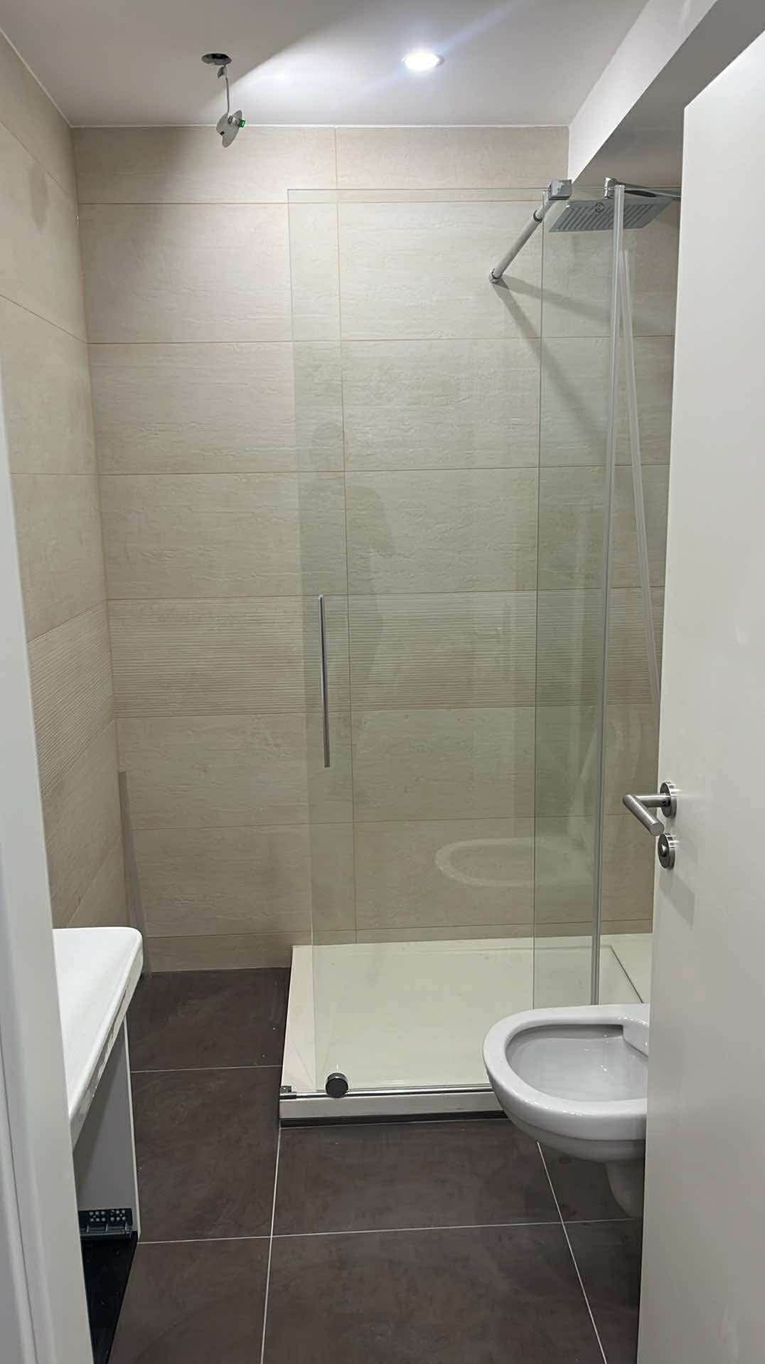 Nowoczesna łazienka po remoncie: kabina prysznicowa ze szklanymi drzwiami, beżowe płytki ścienne z delikatnym wzorem, brązowa podłoga i biała armatura.