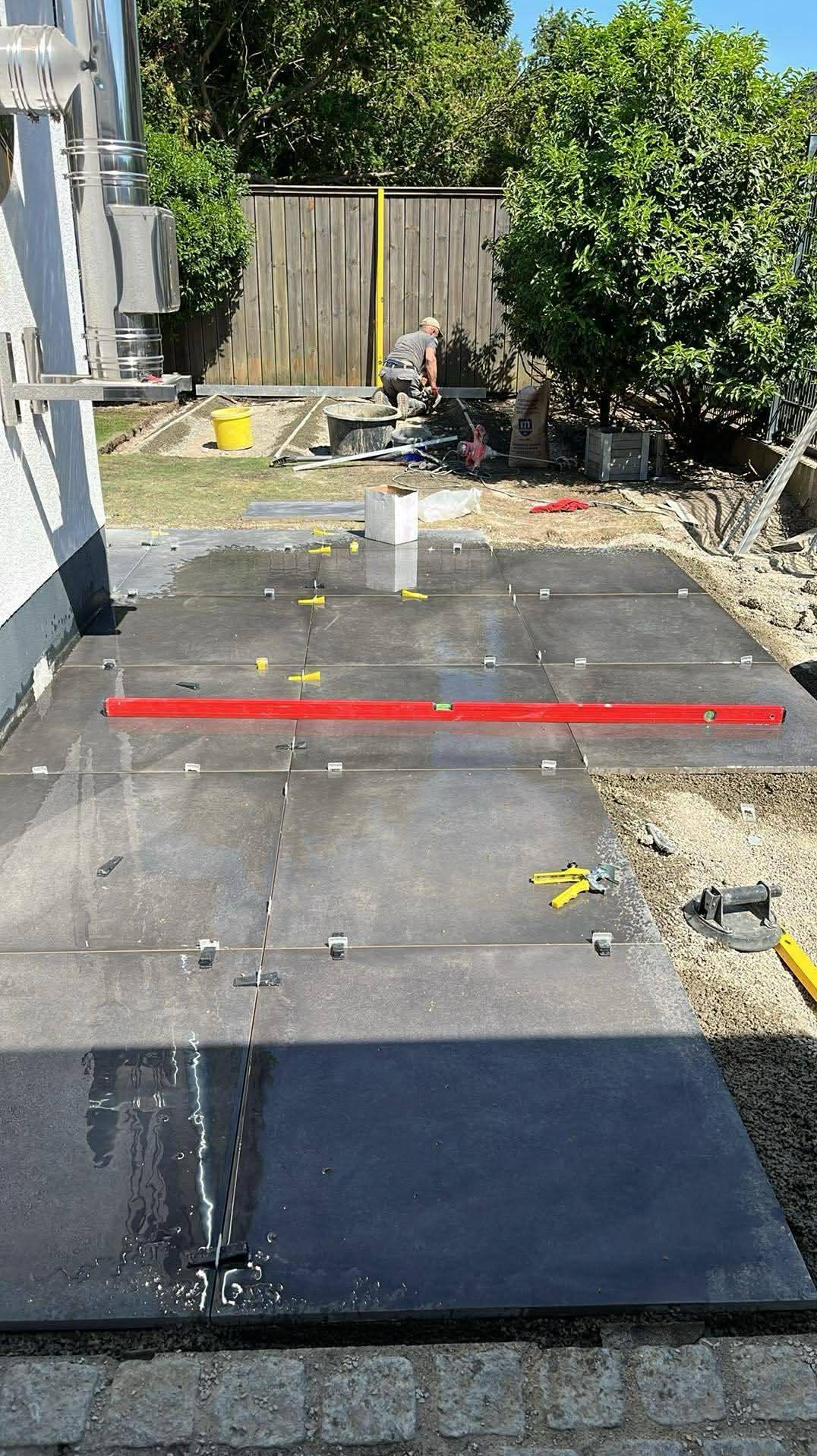 Układanie ciemnych, kwadratowych płyt na zewnątrz budynku. Widoczny poziomica, narzędzia i pracownik w tle. Etap prac w słoneczny dzień.