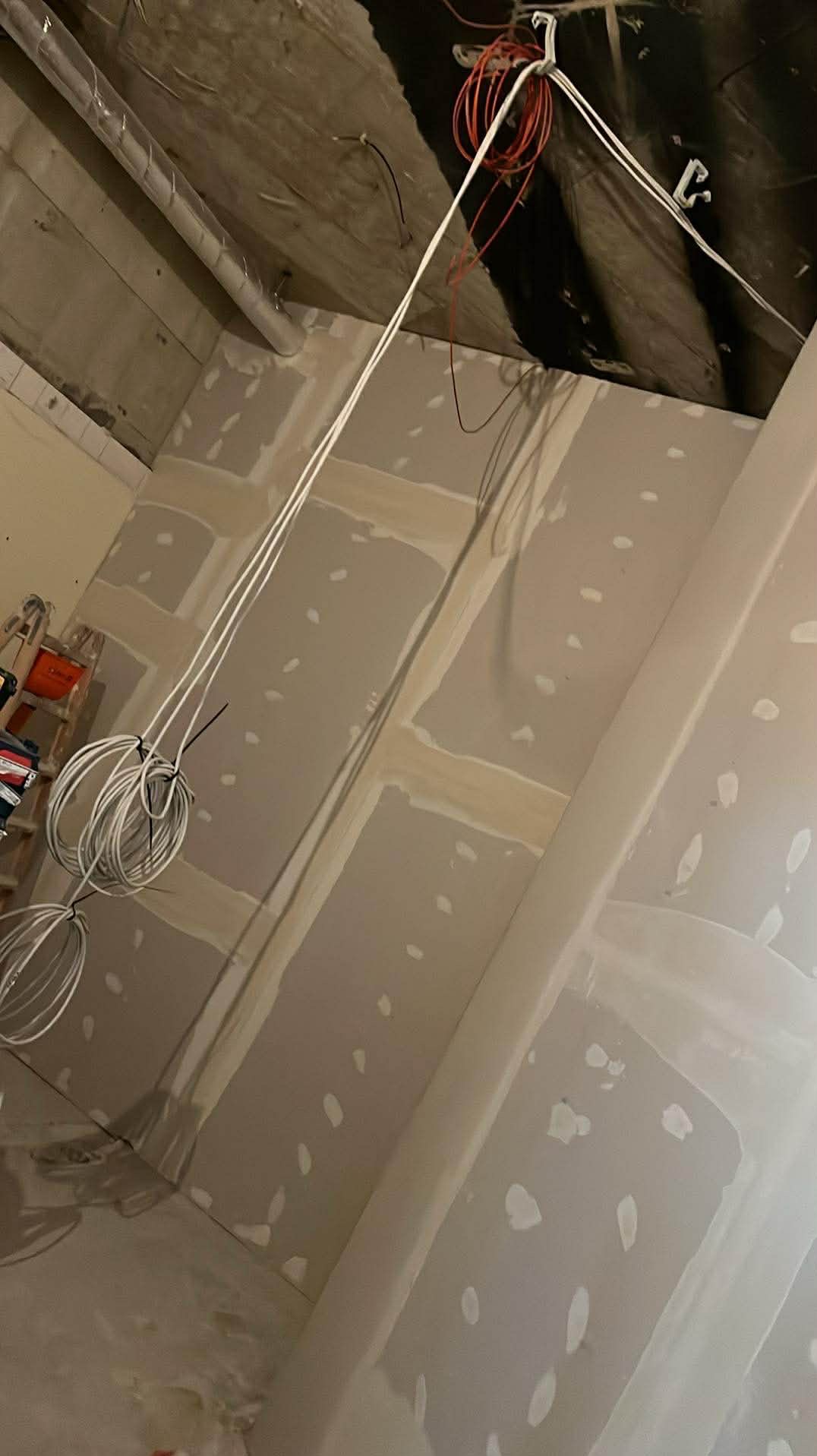 Poddasze w trakcie wykańczania: płyty gipsowo-kartonowe na ścianach i suficie, widoczne łączenia szpachlowane, przewody elektryczne zwisające z sufitu, drabina w tle.