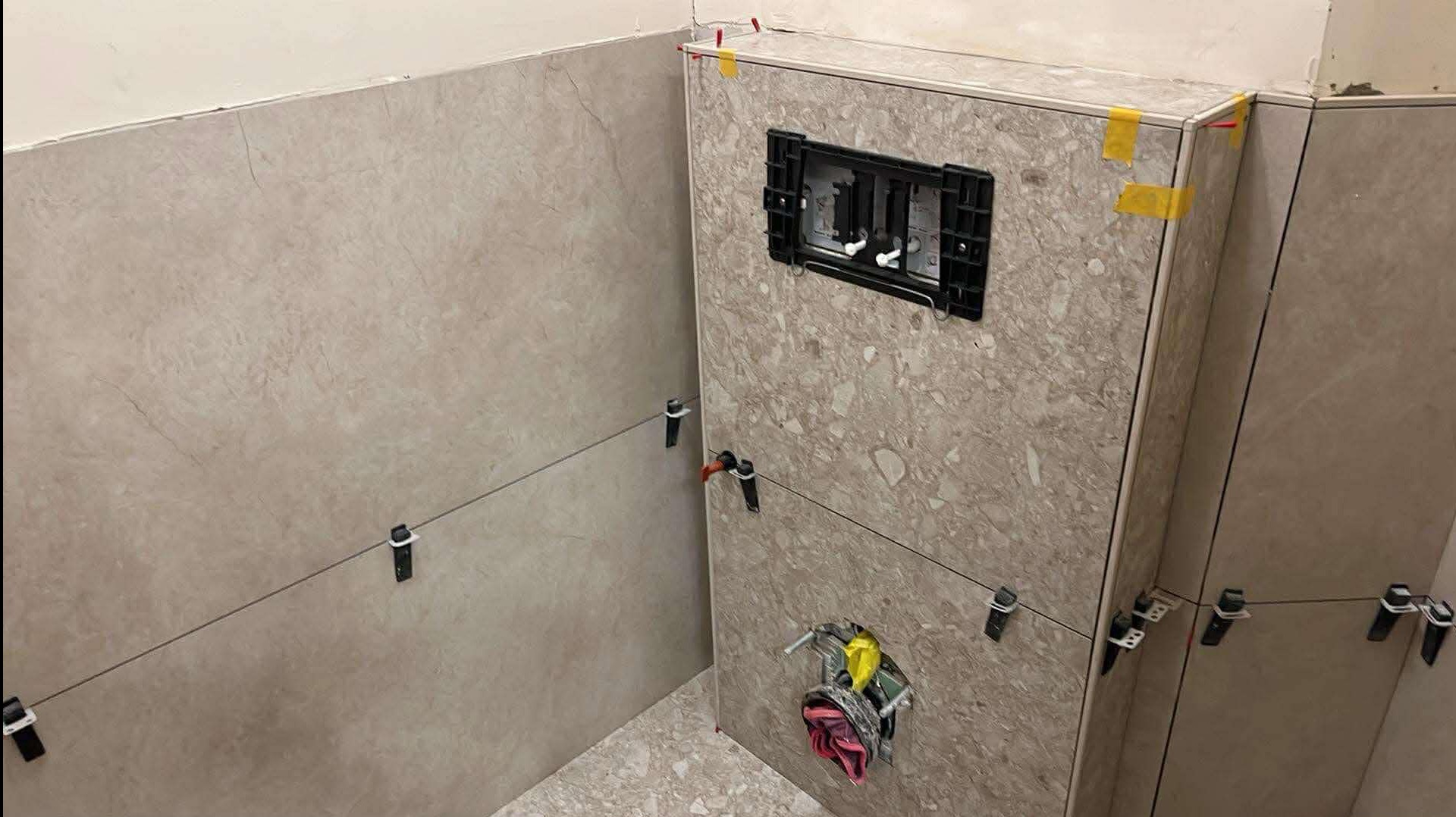 Montaż płytek imitujących marmur w łazience, widoczne narzędzia do poziomowania, instalacja podtynkowa WC, neutralna kolorystyka, etap prac wykończeniowych.