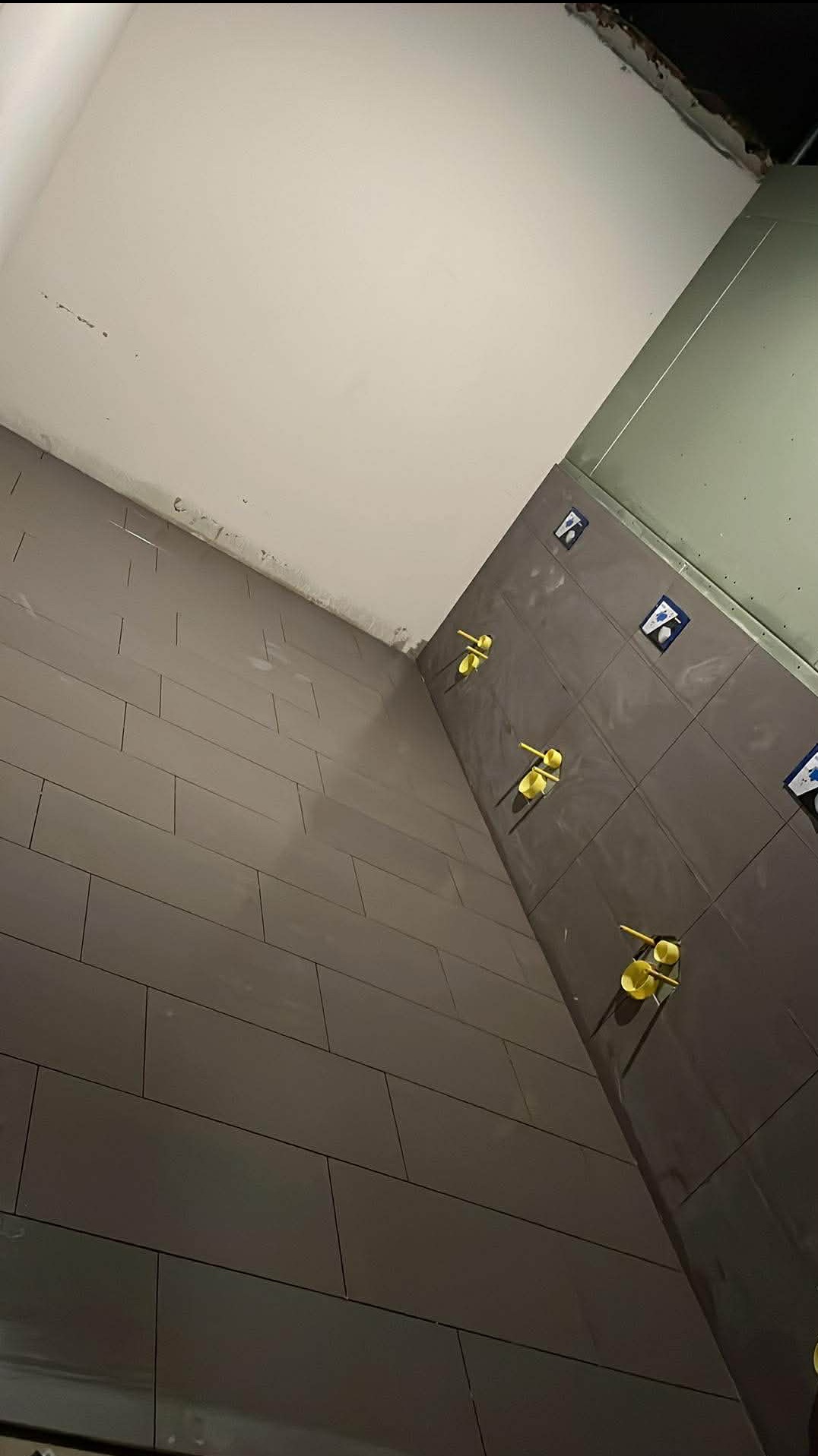 Ukończona ściana łazienki z ciemnymi, prostokątnymi płytkami, z żółtymi krzyżykami dystansowymi, gotowa do fugowania. Widok pod kątem.