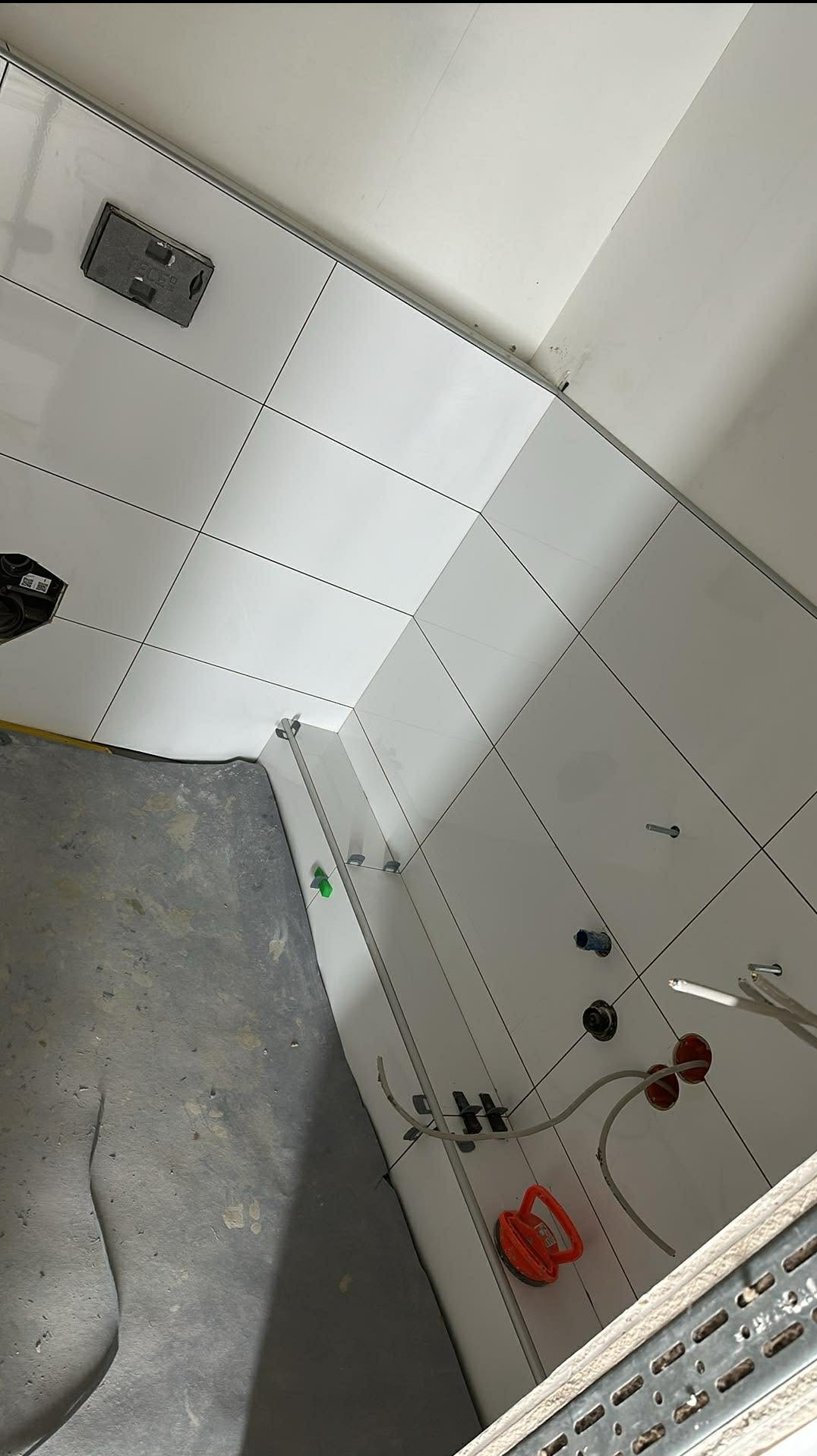 Białe płytki ścienne w trakcie montażu w łazience, widoczne narzędzia glazurnicze i elementy instalacji. Surowy beton na podłodze. Ujęcie z góry, ukośne.