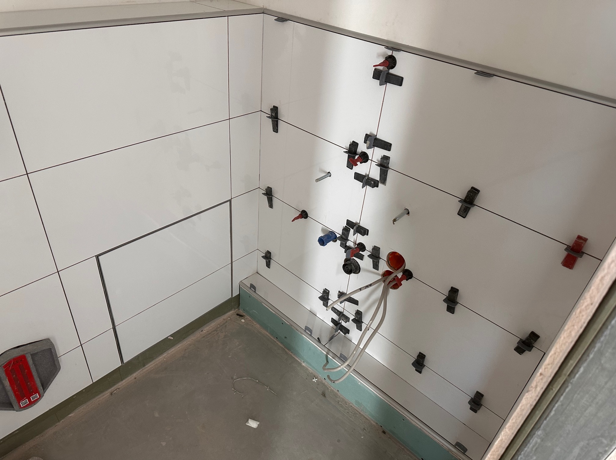 Białe, błyszczące płytki ścienne w trakcie montażu, z użyciem klipsów poziomujących. Widoczne wystające rury instalacji wodnej i fragment podłogi w surowym stanie.