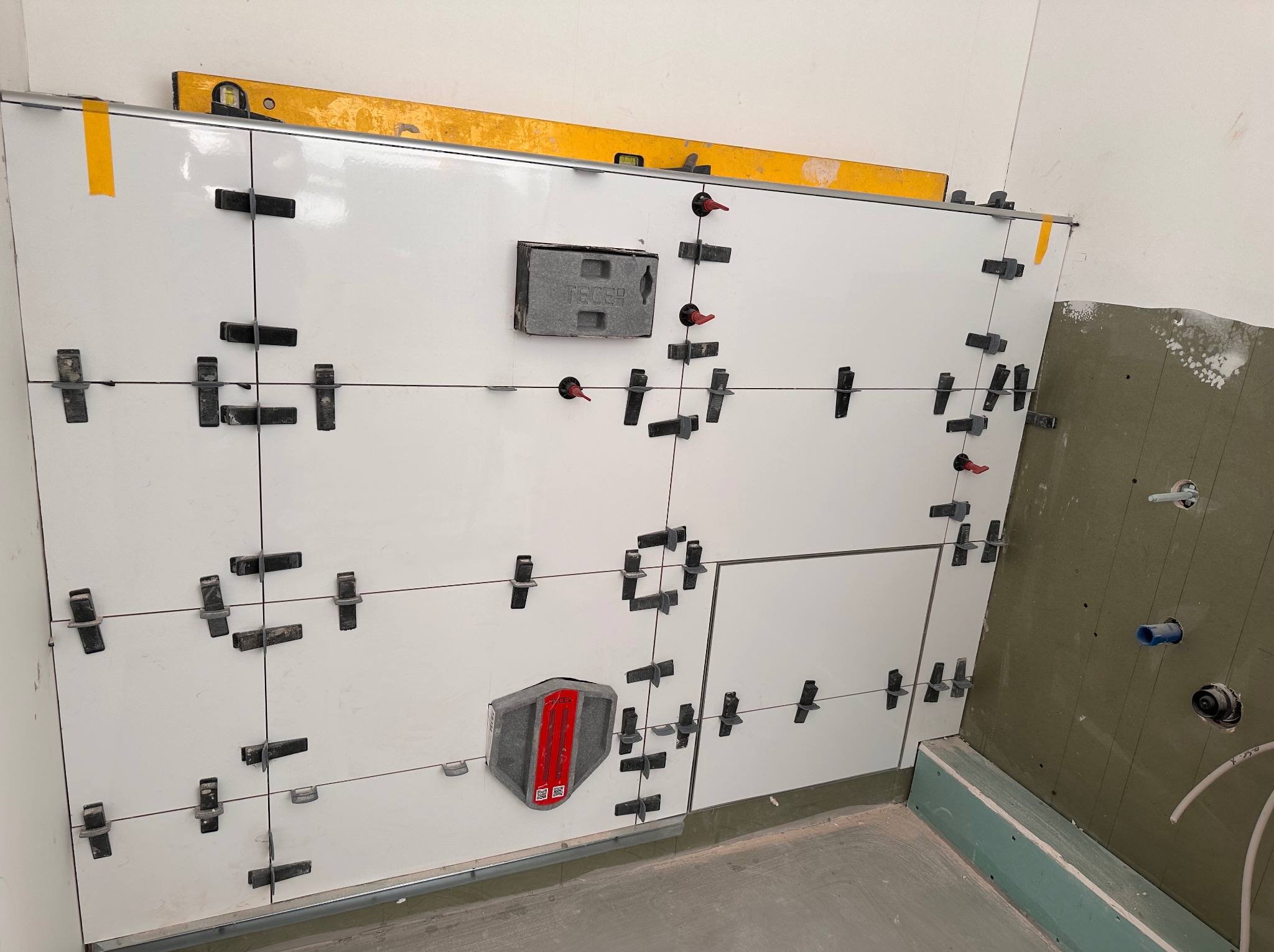 Białe płytki ścienne w trakcie montażu, z użyciem klipsów poziomujących i poziomicy, widoczne elementy instalacji wodnej w surowej łazience.