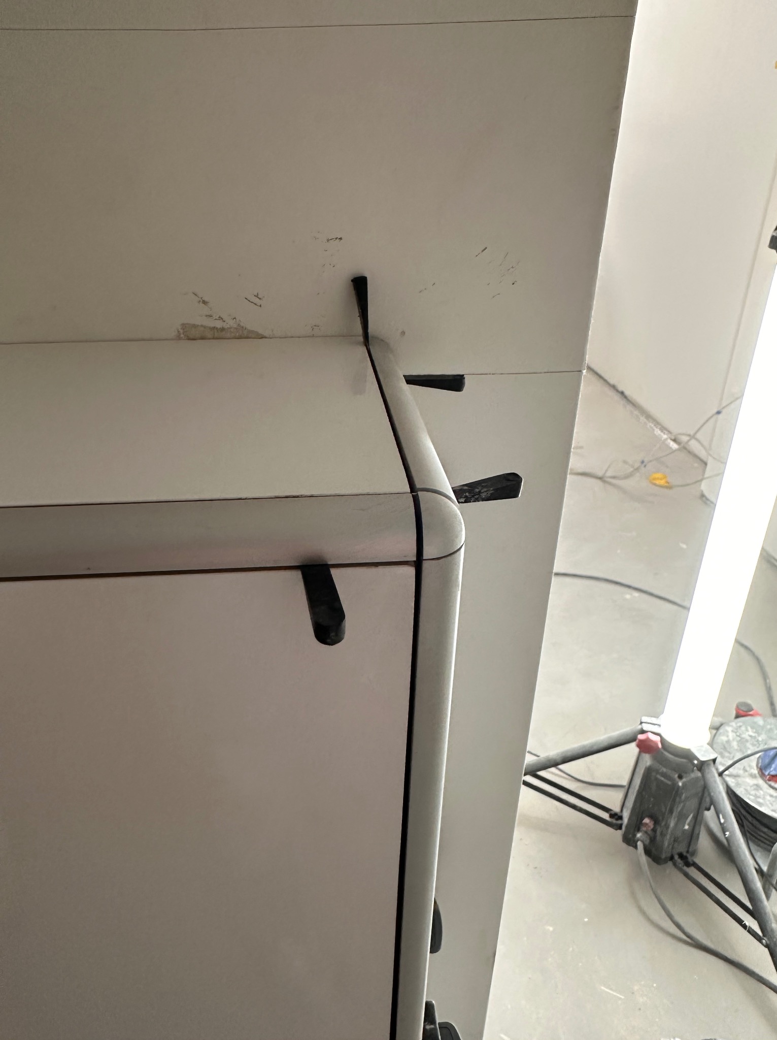 Fragment białej ściany z widocznymi śladami po szpachlowaniu i mocowaniach, oświetlonej lampą budowlaną. Detal wykończenia wnętrza w trakcie prac.