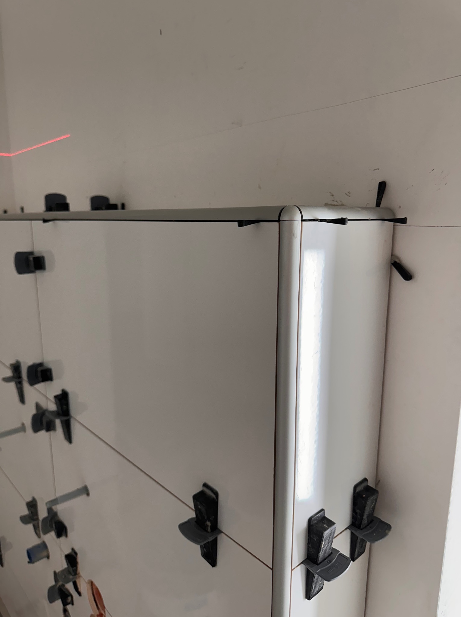 Białe płytki w rogu łazienki, wyrównane za pomocą klipsów i lasera, ukazujące precyzję montażu. Widoczne detale wykończenia i narzędzia glazurnicze.