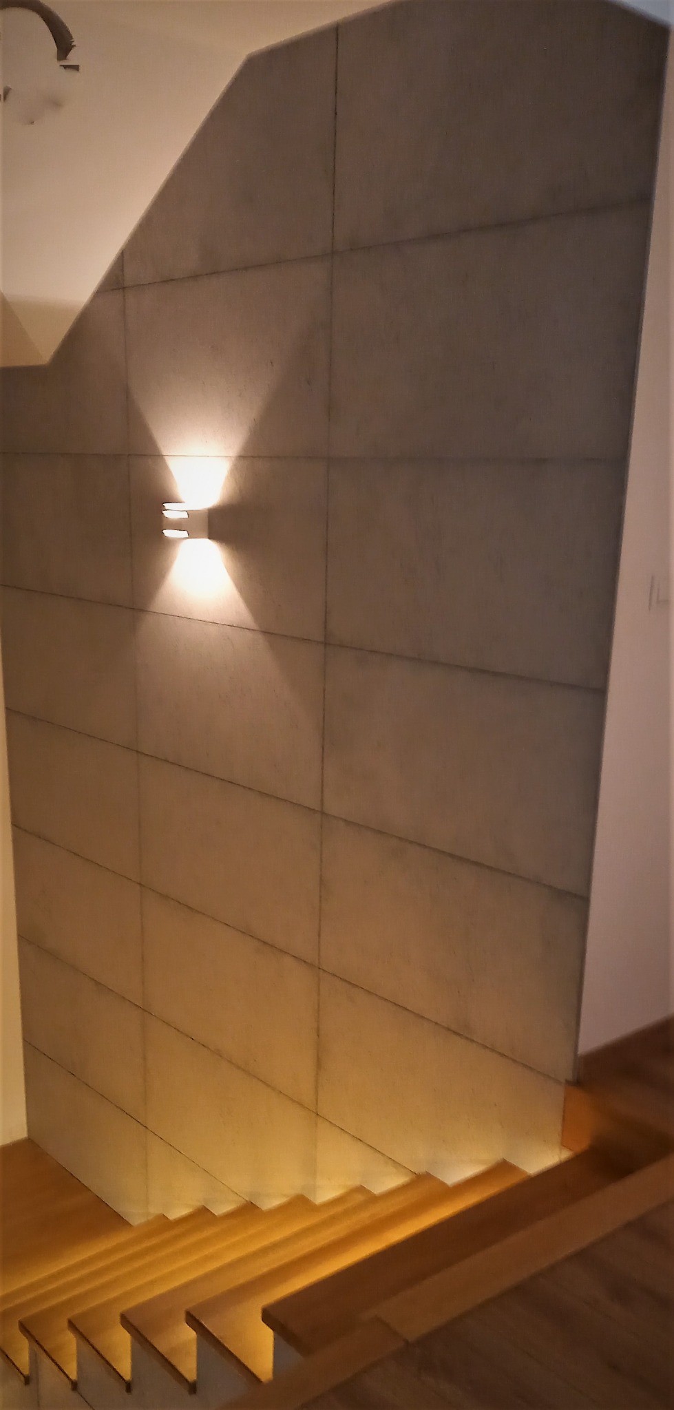 Ściana w klatce schodowej wyłożona panelami imitującymi beton, oświetlona kinkietem i podświetlonymi stopniami schodów.