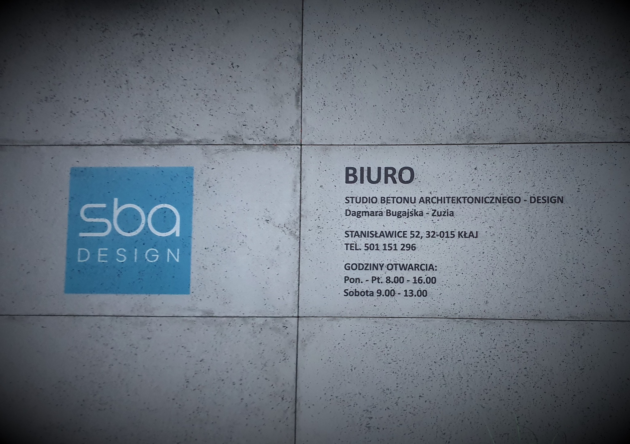 Tablica informacyjna z nazwą i danymi kontaktowymi Studia Betonu Architektonicznego Design SBA na szarym, betonowym tle.