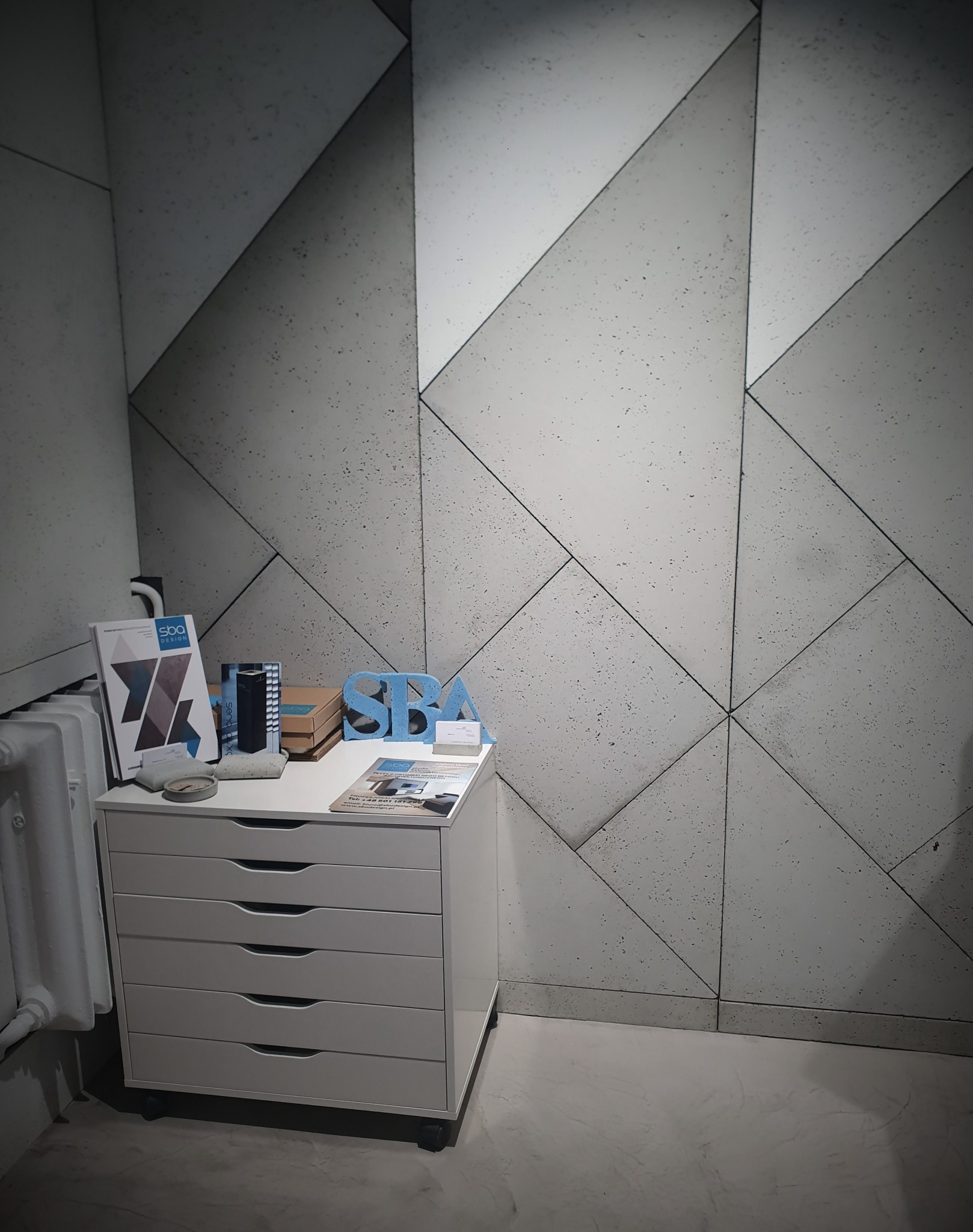 Ściana wyłożona panelami w geometryczne wzory z betonu architektonicznego, z bliska, z widoczną szafką z materiałami reklamowymi.