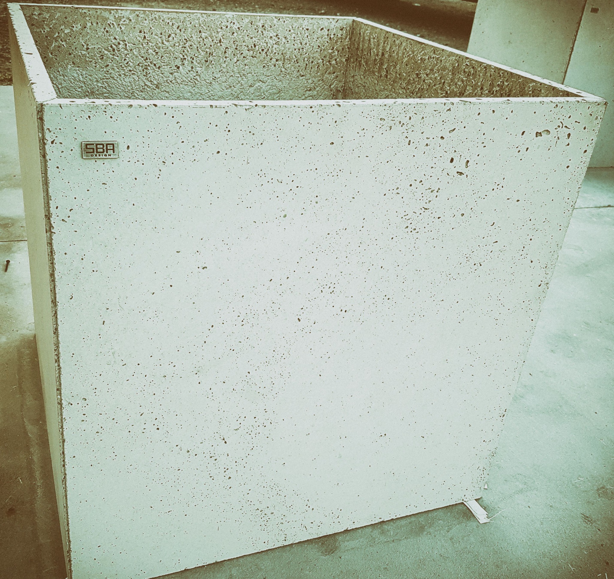 Kwadratowa donica z betonu architektonicznego o porowatej strukturze, z widocznym logo producenta, ustawiona na zewnątrz.