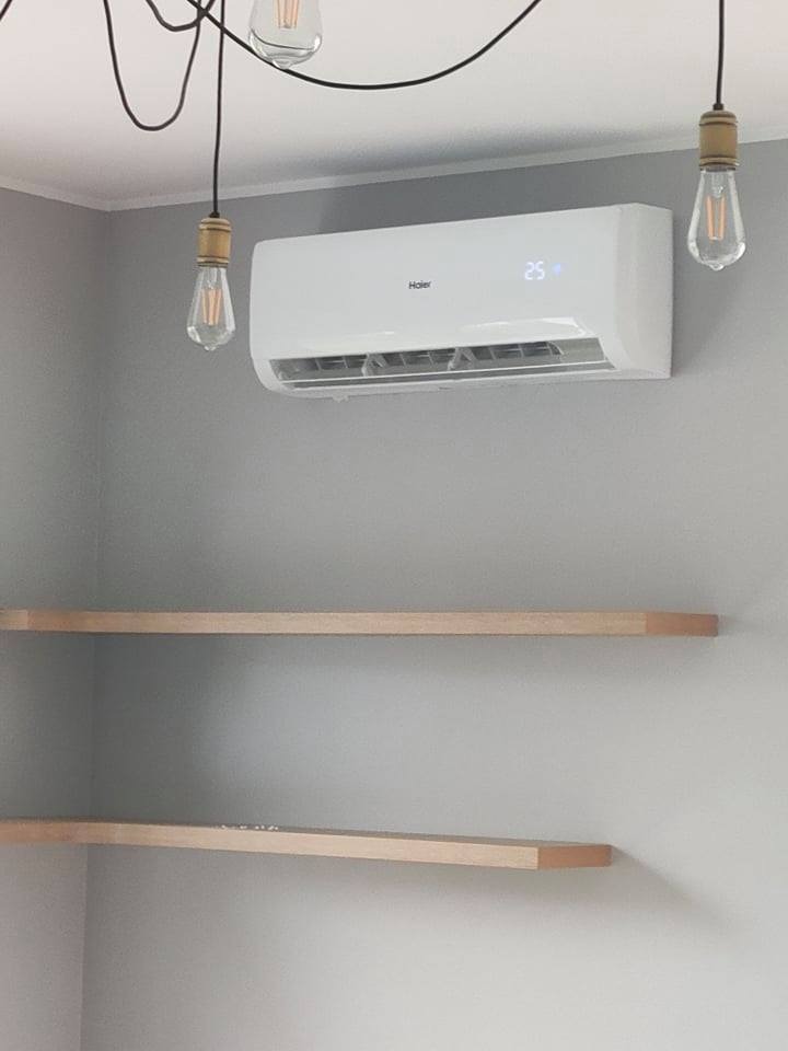 Biała klimatyzacja Haier na szarej ścianie, z wyświetlaczem pokazującym 25 stopni, oświetlona przez dwie wiszące żarówki Edisona i z dwoma drewnianymi półkami poniżej.