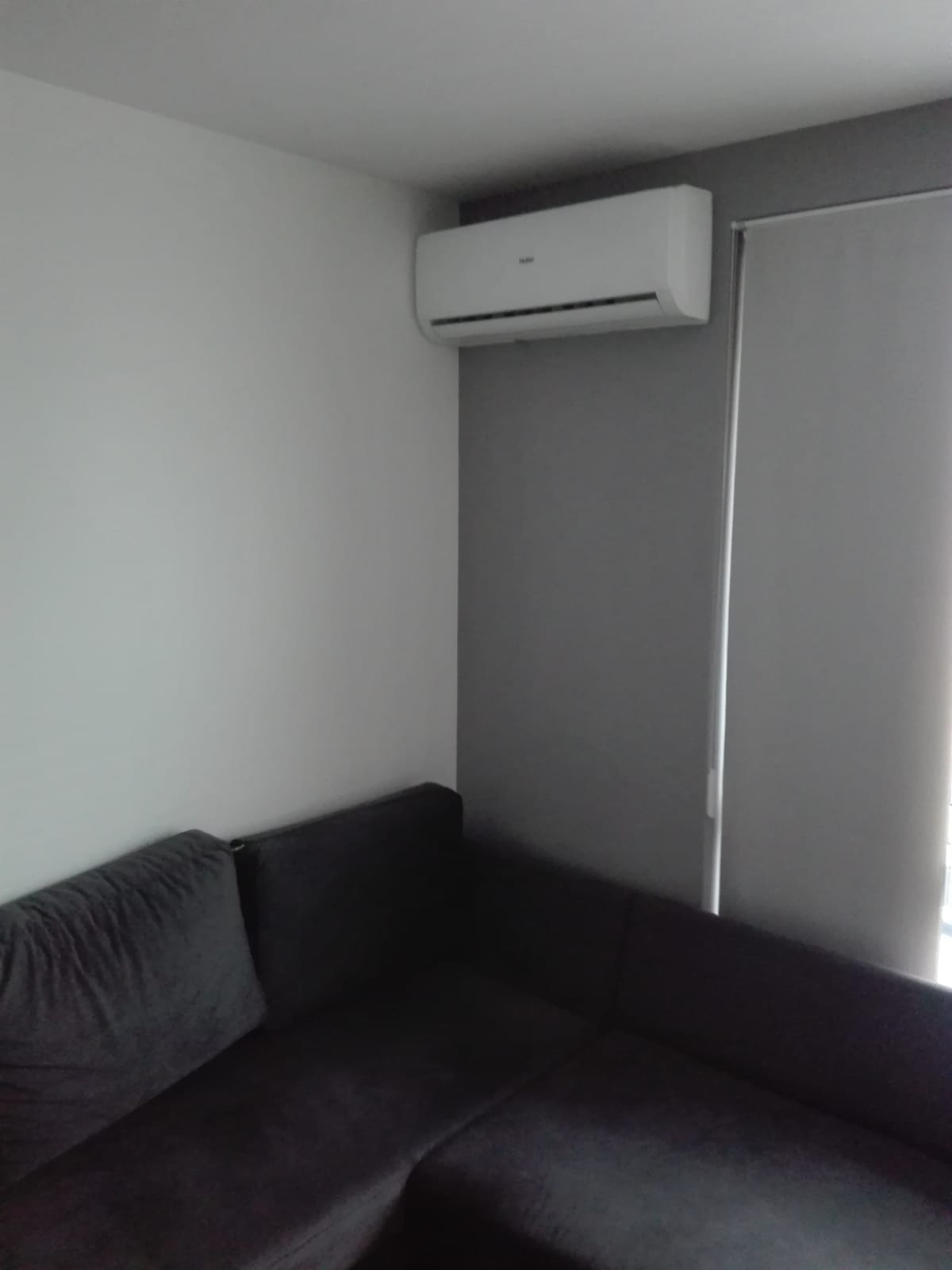 Biała jednostka klimatyzacji Haier zamontowana na szarej ścianie w rogu pokoju nad ciemnoszarą sofą narożną, z widoczną roletą okienną po prawej stronie.