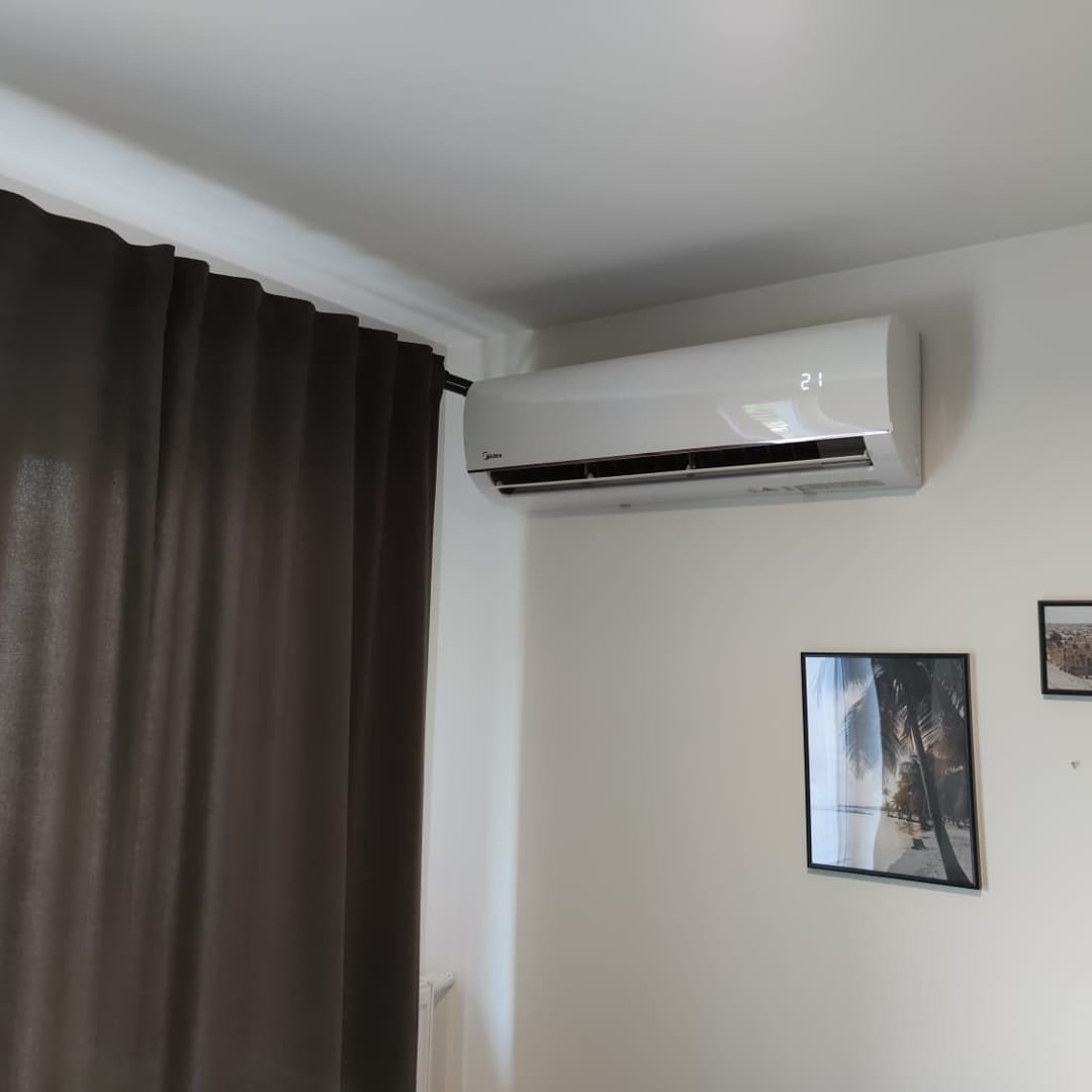 Biały klimatyzator Midea zamontowany na ścianie w pokoju z ciemnymi zasłonami i obrazkami w ramkach.
