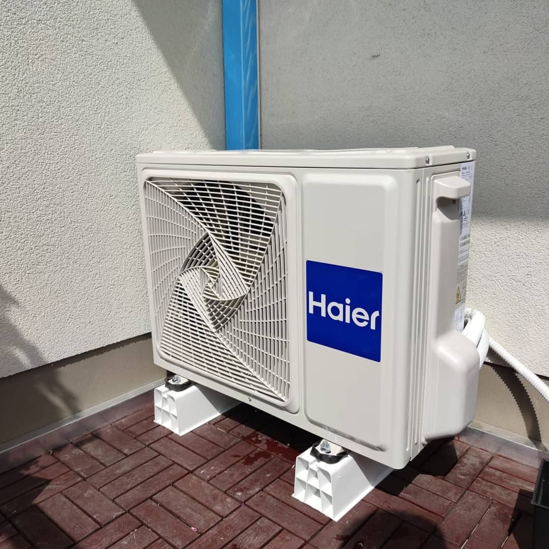 Zewnętrzna jednostka klimatyzacji Haier zamontowana na białych wspornikach na brązowej, ceglanej podłodze, przylegająca do ściany budynku.