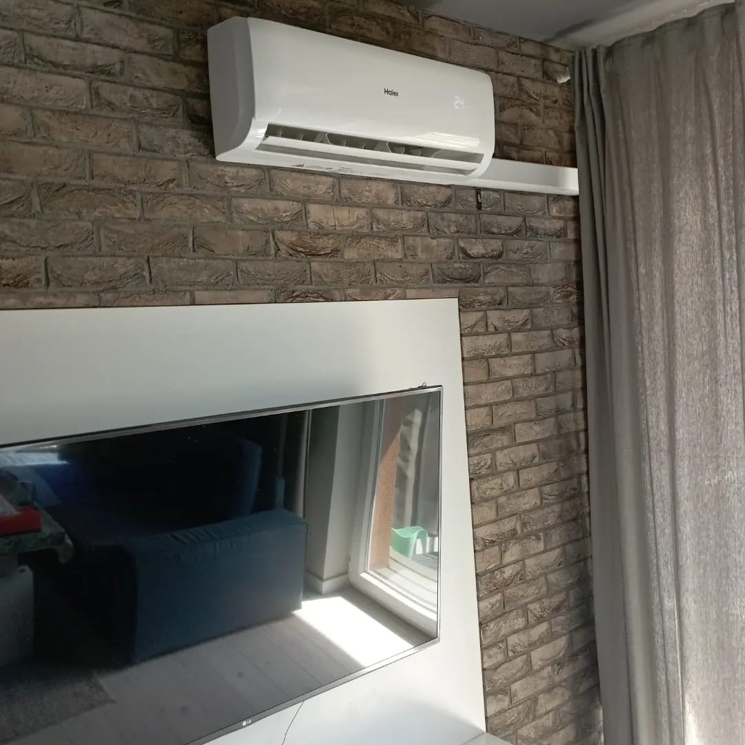 Biały klimatyzator Haier zamontowany na ścianie z cegły w pokoju z zasłoniętym oknem i odbiciem wnętrza w ekranie telewizora.