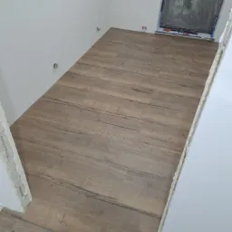 Ułożona podłoga z paneli imitujących drewno w jasnym wnętrzu, okno zabezpieczone folią malarską, widoczne gniazdka elektryczne bez osłon.