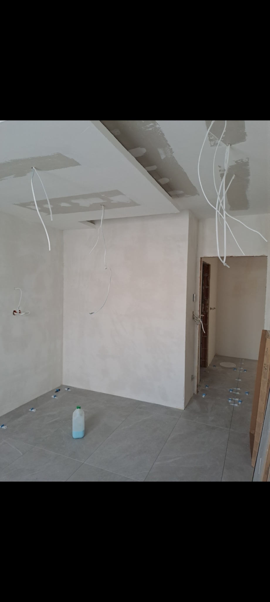 Wnętrze w trakcie prac w Rudzie Śląskiej: surowe ściany po szpachlowaniu, wystające przewody elektryczne z sufitu podwieszanego i świeżo położone szare płytki podłogowe z niebieskimi krzyżykami...