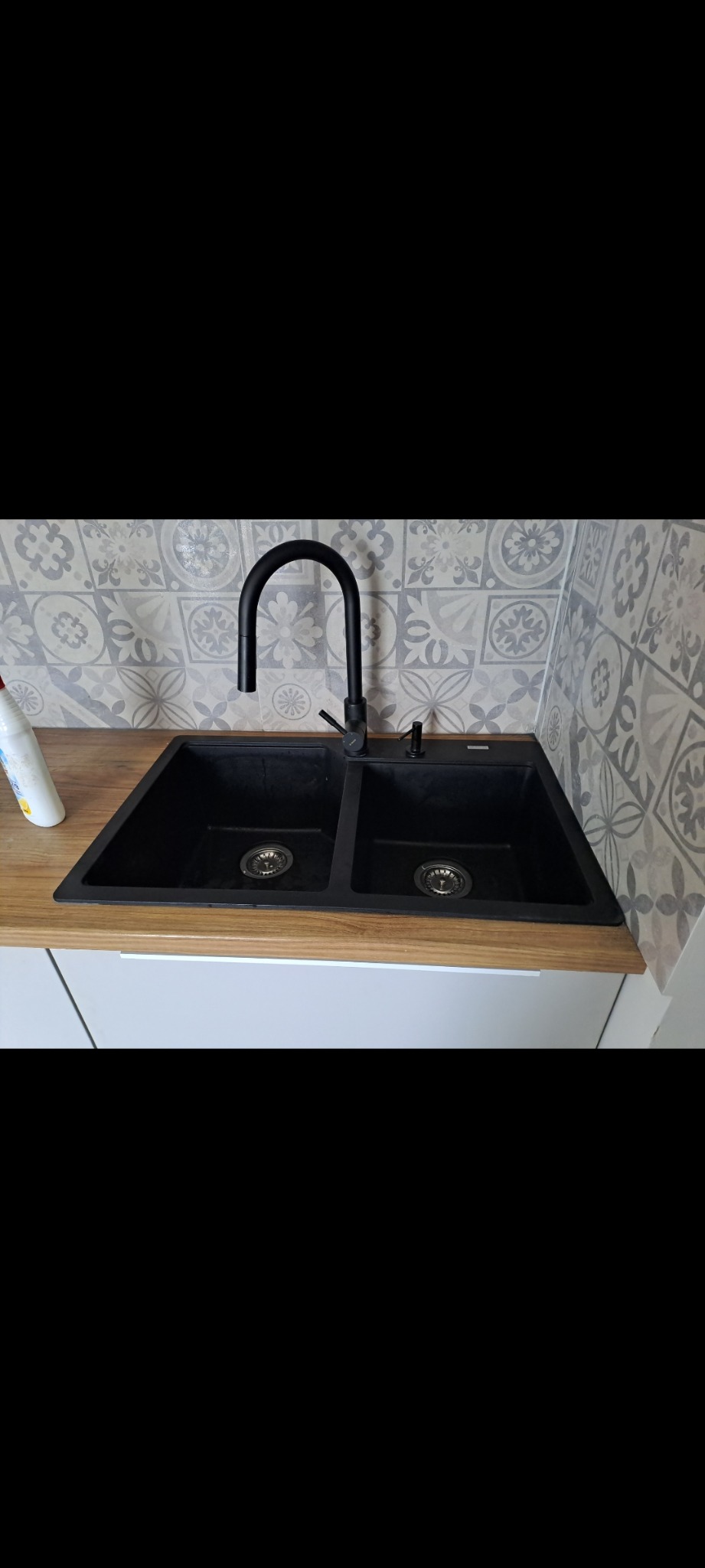 Czarny, dwukomorowy zlew granitowy z czarną baterią, zamontowany w jasnym blacie drewnopodobnym, na tle ściany z dekoracyjnymi, szarymi płytkami.