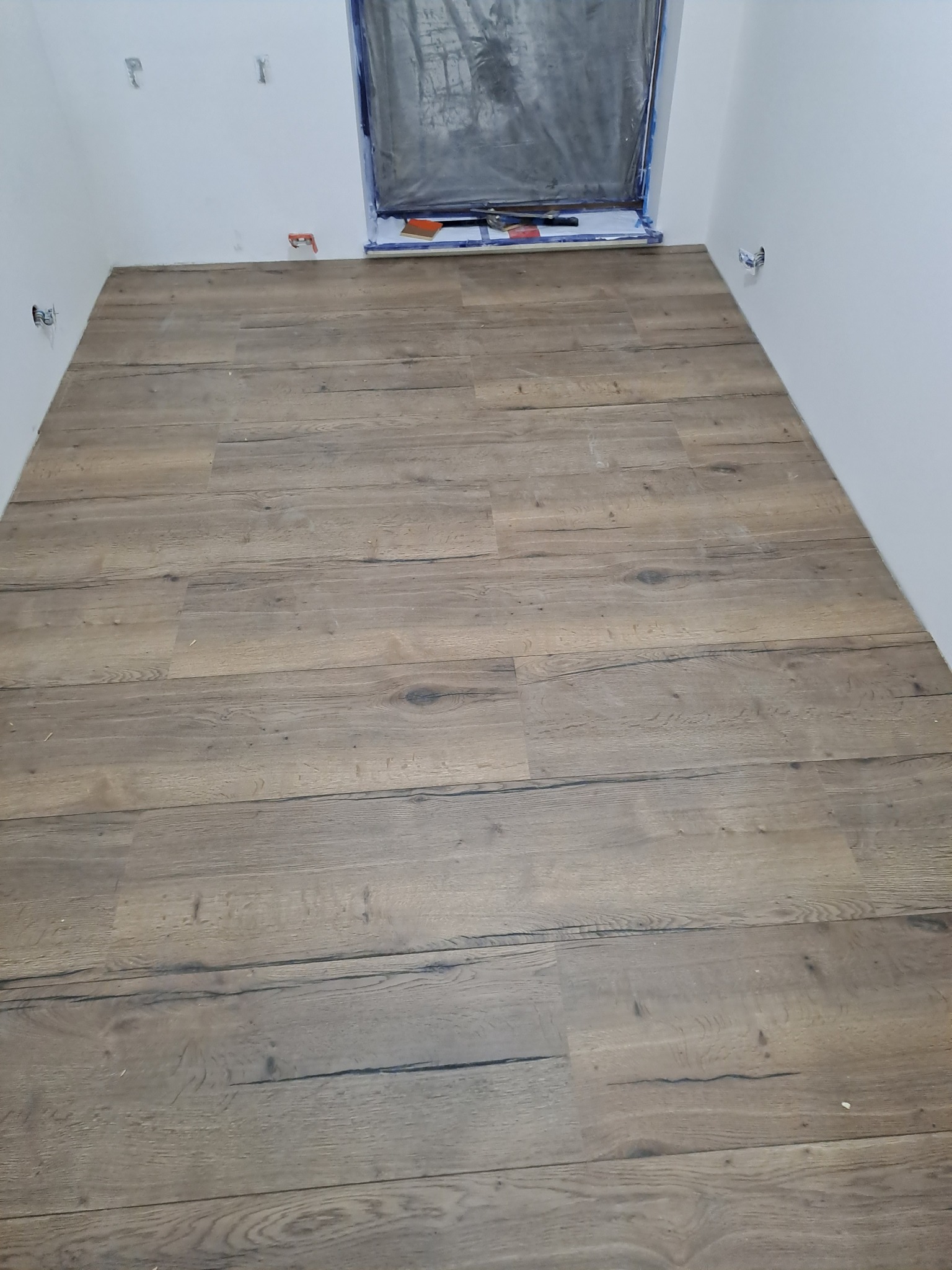 Nowo ułożona podłoga z paneli imitujących stare drewno, widoczne listwy przypodłogowe jeszcze nie zamontowane, okno zabezpieczone folią malarską.