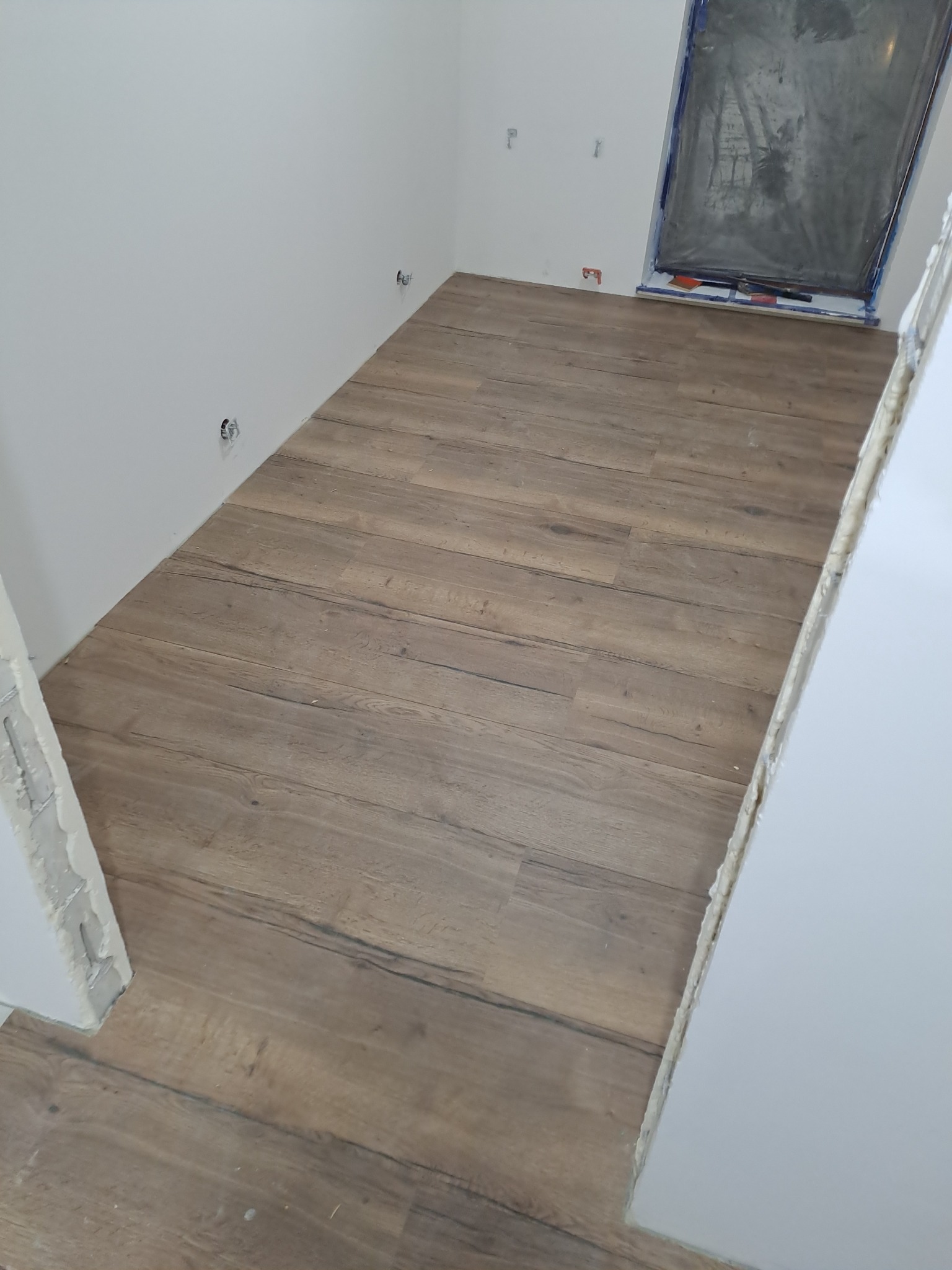 Ułożona podłoga z paneli imitujących drewno w jasnym wnętrzu, okno zabezpieczone folią malarską, widoczne gniazdka elektryczne bez osłon.