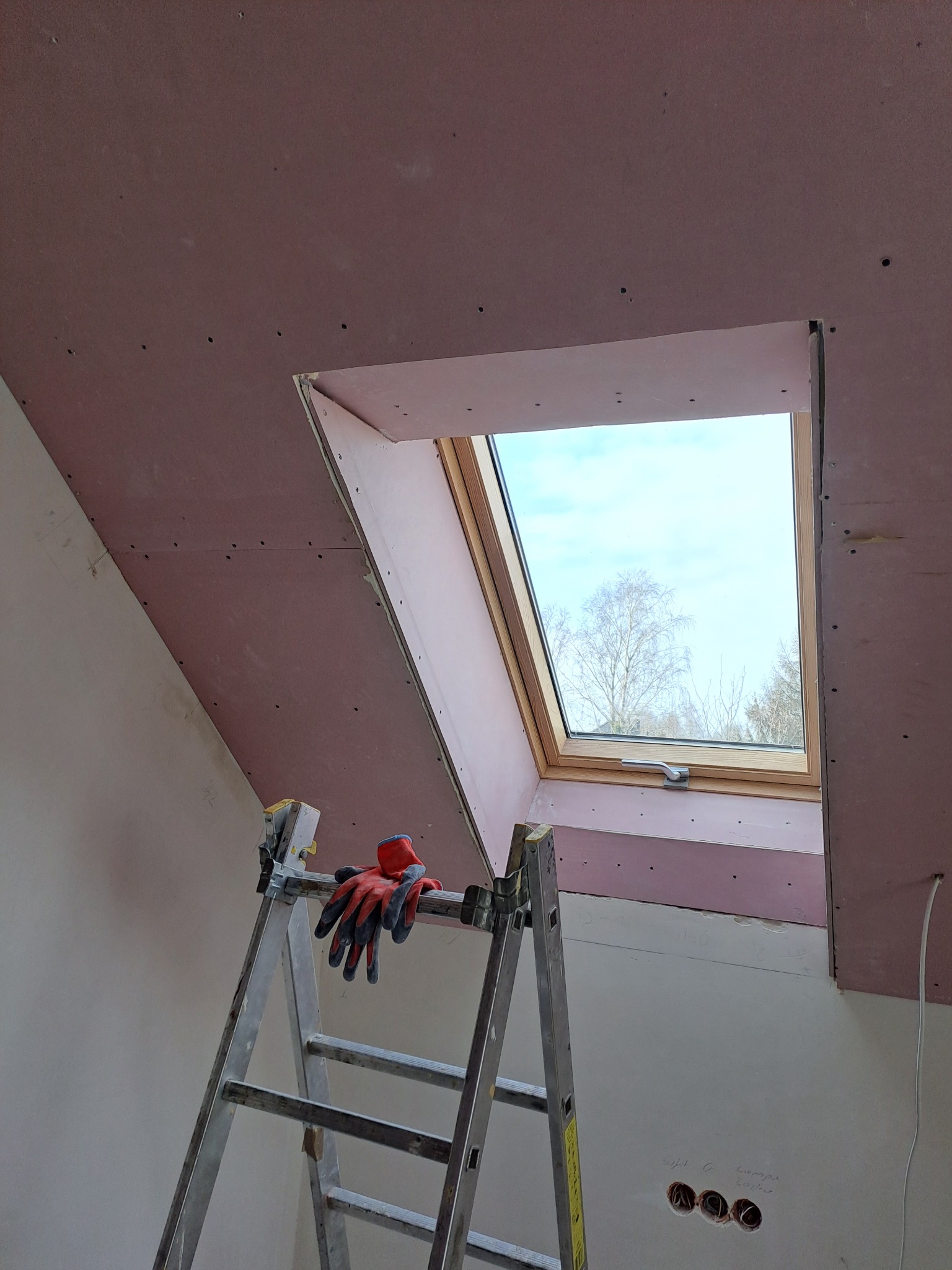 Montaż okna dachowego w poddaszu, widoczne płyty gipsowo-kartonowe na skosach, drabina z rękawicami roboczymi i otwory na gniazdka elektryczne w ścianie.