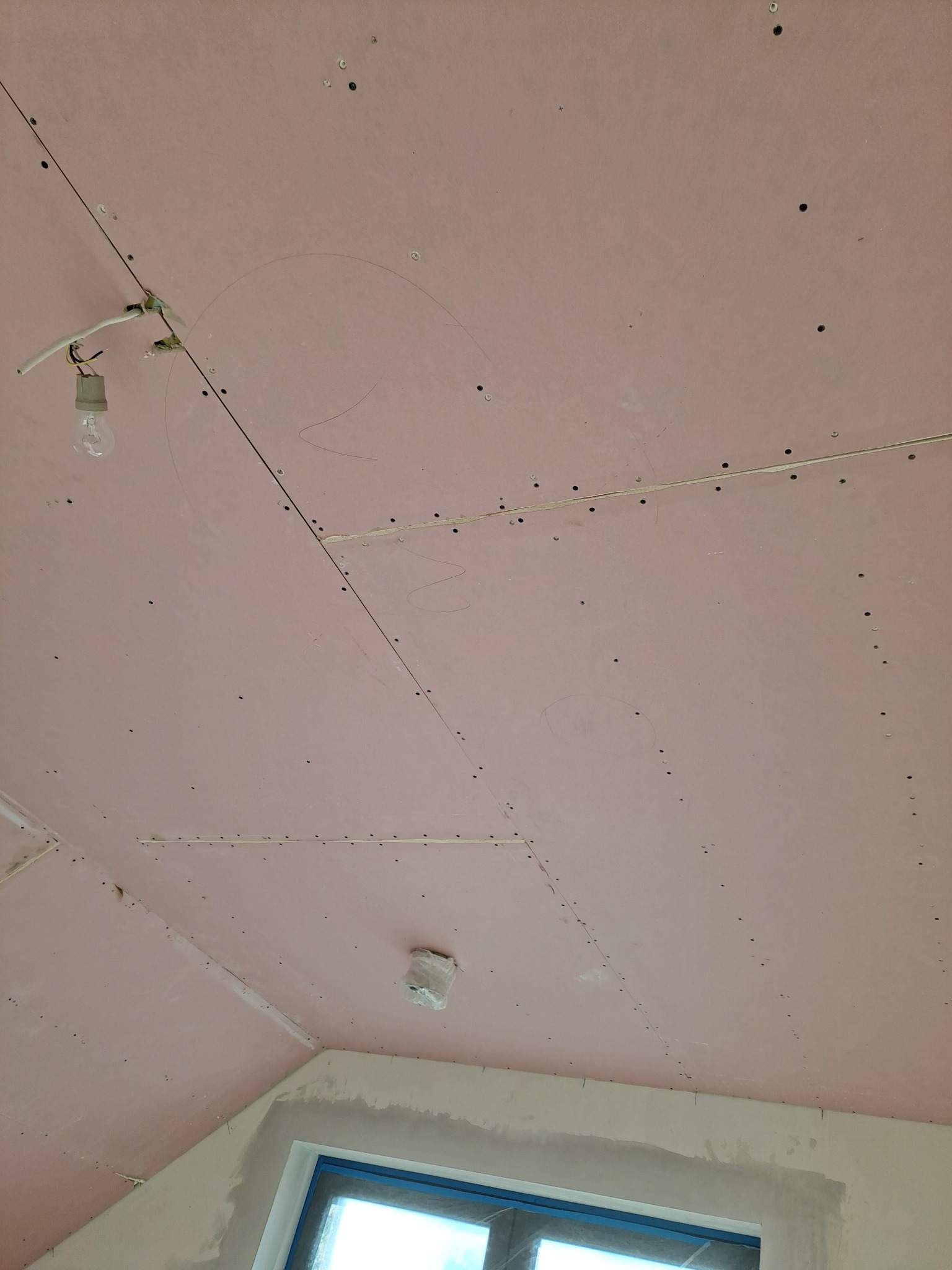 Różowa płyta gipsowo-kartonowa na suficie poddasza, widoczne ślady ołówka, kabel z żarówką i otwory po wkrętach, okno w dolnej części kadru.