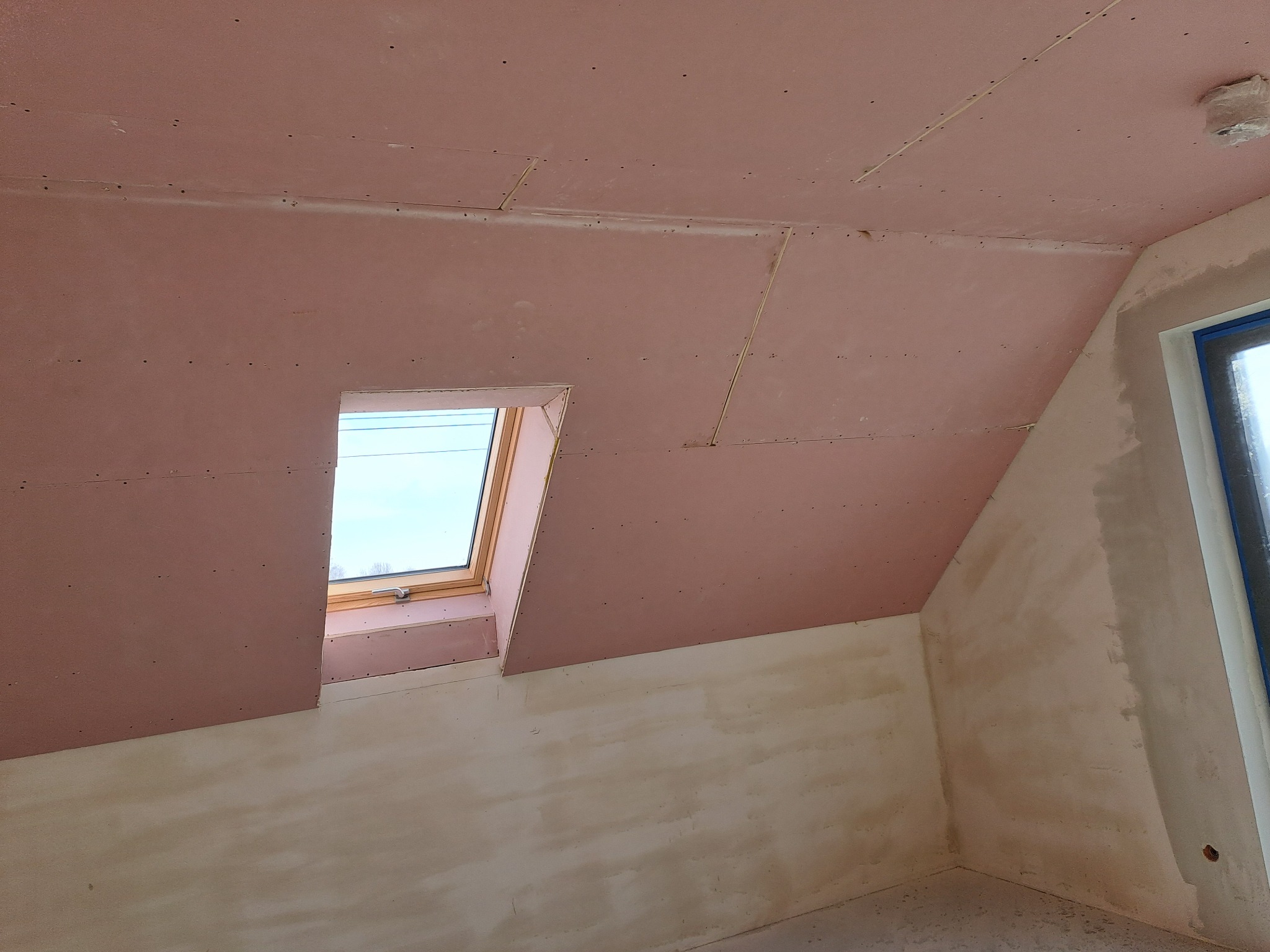 Poddasze z oknem dachowym w trakcie prac wykończeniowych: płyty gipsowo-kartonowe na skosach sufitu i ścianach, widoczne łączenia płyt przed szpachlowaniem, ściany częściowo pokryte warstwą gładzi.