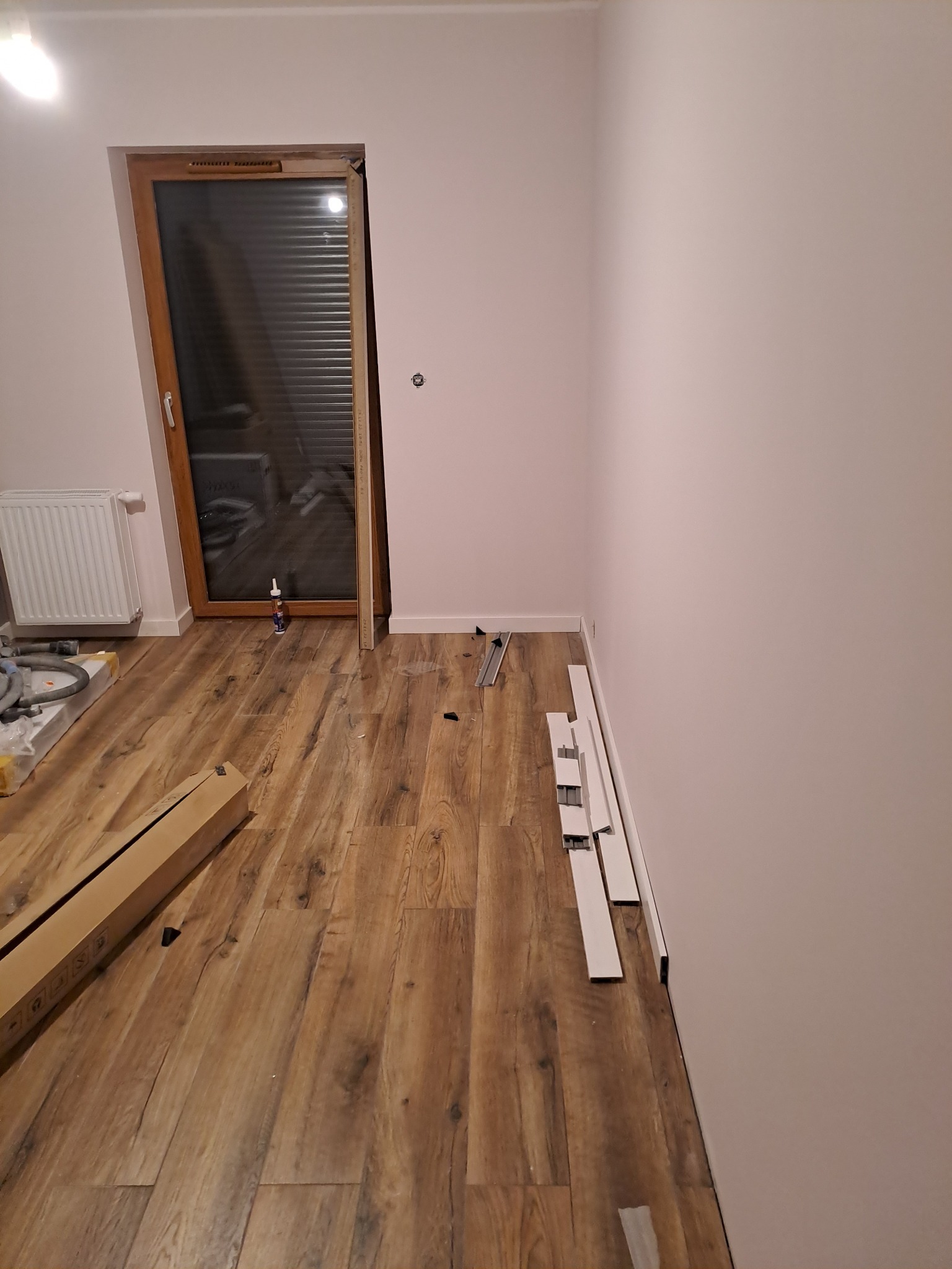 Pokój w trakcie prac, widoczne panele podłogowe imitujące drewno, białe listwy przypodłogowe ułożone wzdłuż ściany, karton z panelami oraz narzędzia w tle