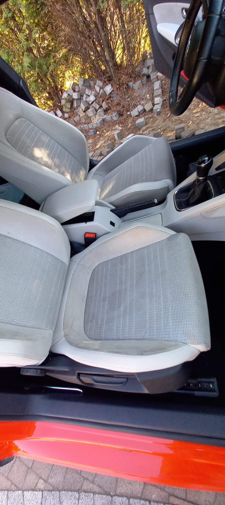 Wnętrze samochodu z jasną tapicerką, widoczne fotele z tkaniny o delikatnym wzorze, z otwartymi drzwiami na tle kostki brukowej i zieleni.