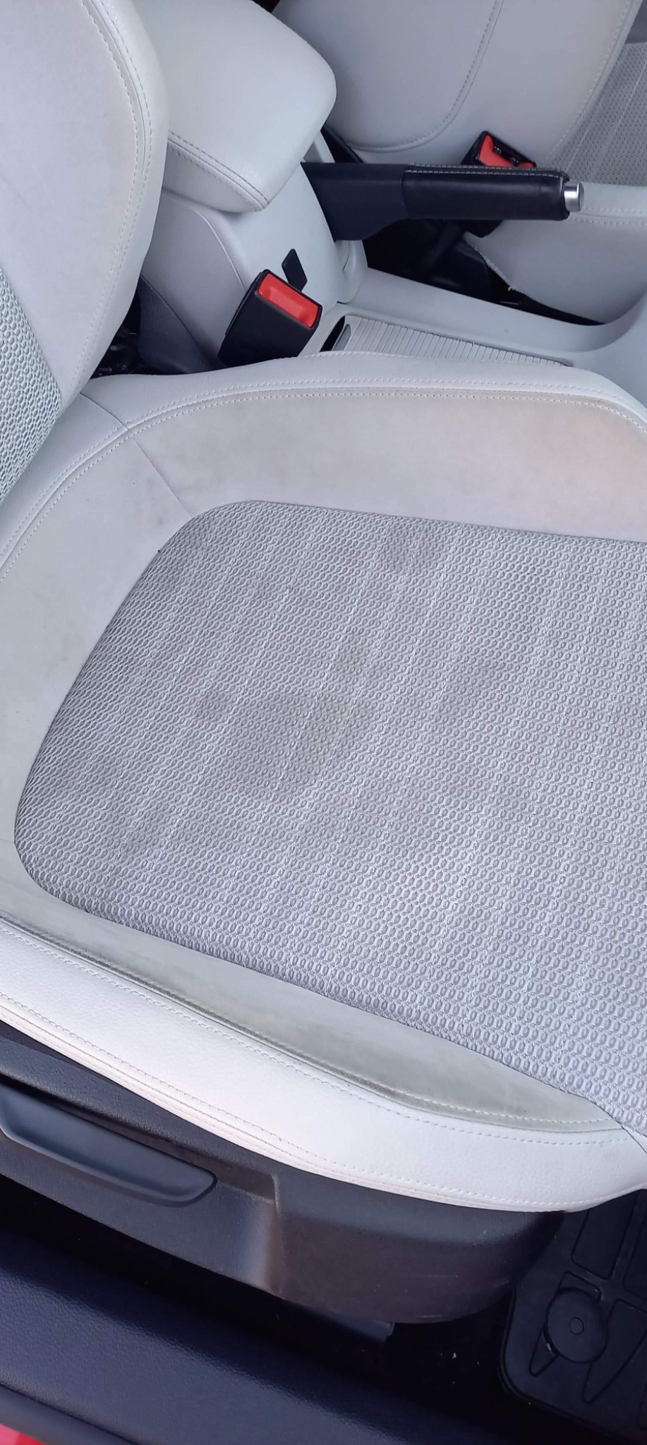 Jasna tapicerka fotela samochodowego z widocznymi zabrudzeniami i plamami, zapięcie pasów bezpieczeństwa w tle.