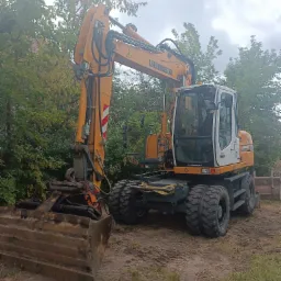 Żółto-biała koparka Liebherr na nierównym terenie z zielenią w tle, widoczny duży łyżka koparki i opony z grubym bieżnikiem.
