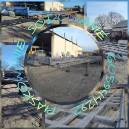 Metalowa rama przyczepy na piasku, po obróbce, widoczne ślady po piaskowaniu, na tle budynki gospodarcze i stare pojazdy.