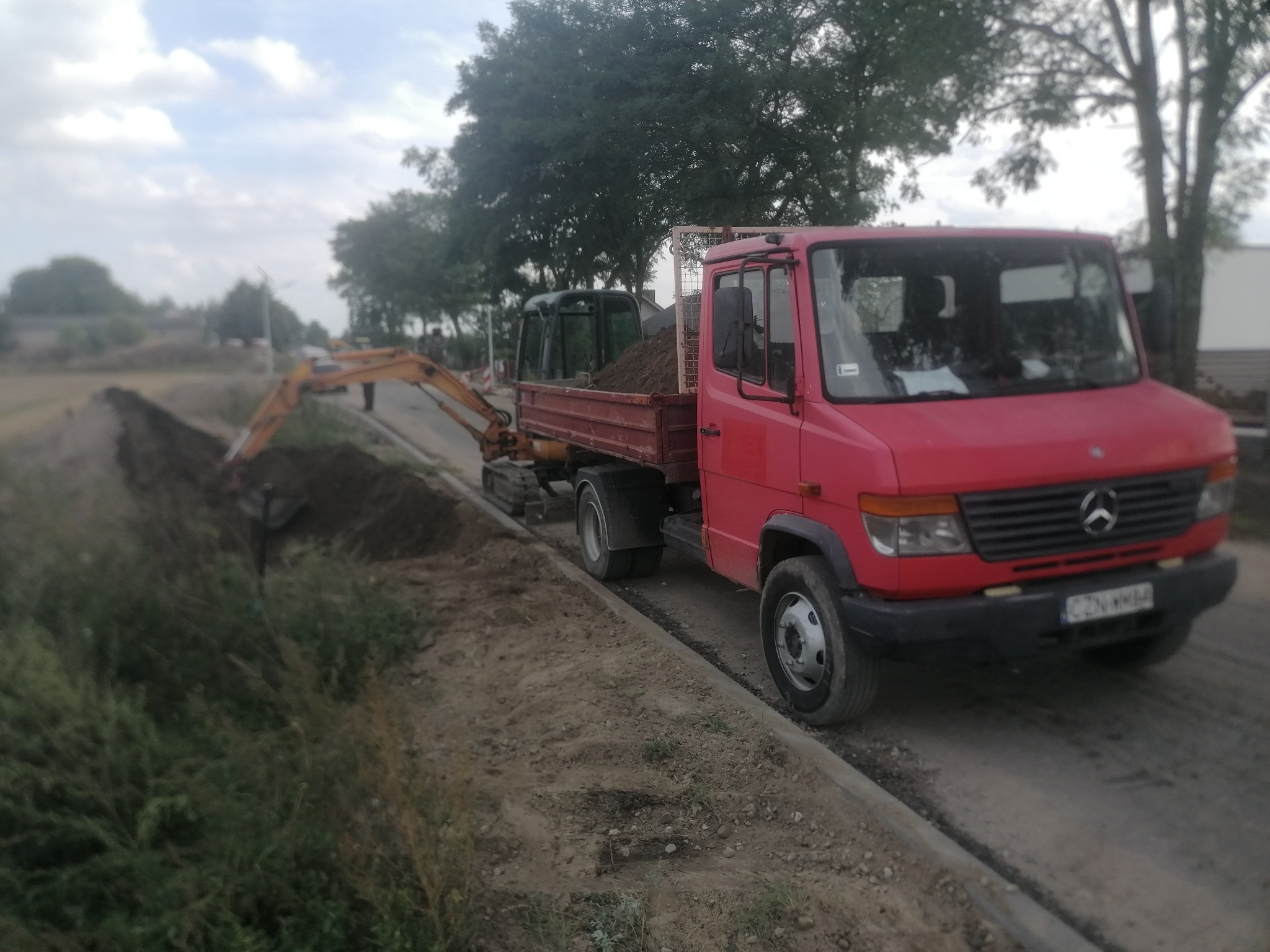 Czerwona ciężarówka z mini koparką na pace, ziemia i wykop w tle. Prace ziemne w toku, widok na wykopany rów i zieleń.