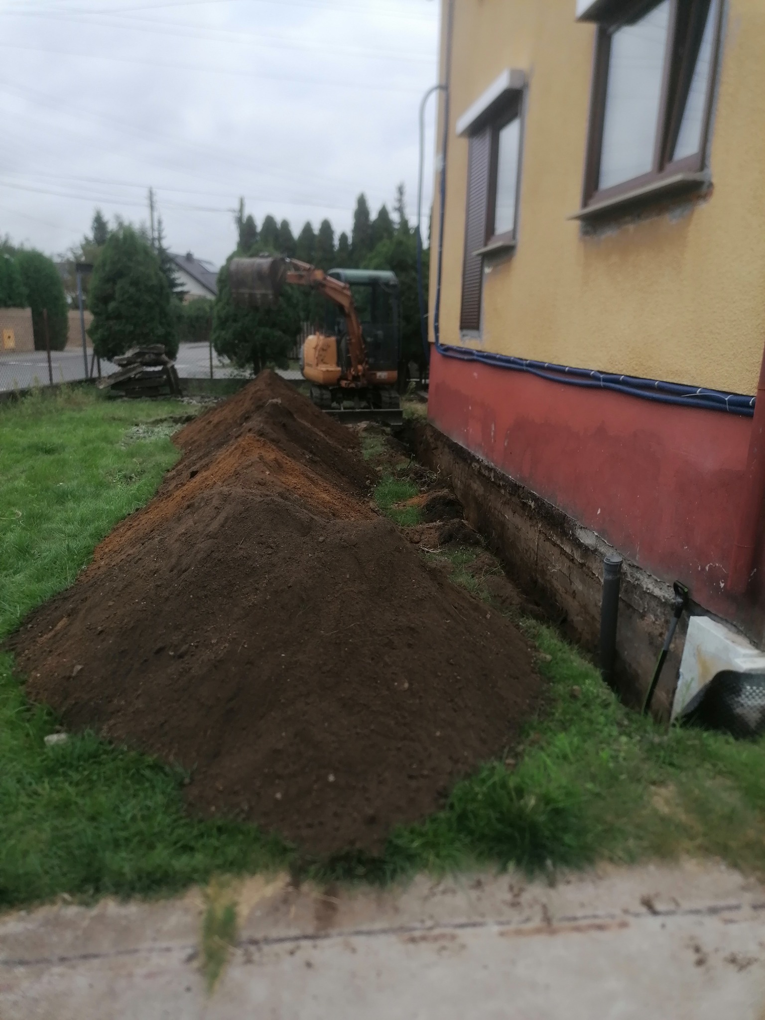 Wykop pod fundamenty domu jednorodzinnego, z widocznym wykopem wzdłuż ściany i stertą ziemi. Na drugim planie mała koparka i budynek z żółtą elewacją.