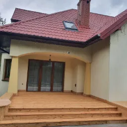 Nowy drewniany taras z trzema stopniami przed wejściem do domu z beżową elewacją i czerwoną dachówką, widok z dołu.