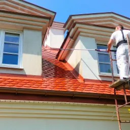 Pracownik w kombinezonie maluje elewację budynku z rusztowania za pomocą długiego wałka. Widoczna pomalowana część dachu i elewacji. Błękitne niebo w tle.