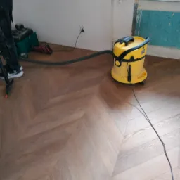 Ułożona, brązowa podłoga z paneli w jodełkę, czyszczona żółtym odkurzaczem przemysłowym; widoczne narzędzia i fragment osoby. Wykończenie podłogi.