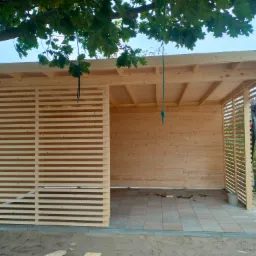 Drewniana altana z poziomymi listwami, częściowo zadaszona, z podłogą wyłożoną kostką, liny zwisające z dachu, widok spod drzewa.