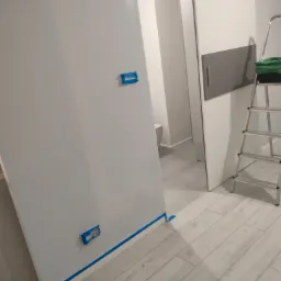 Świeżo pomalowana, jasnoszara ściana z niebieską taśmą malarską zabezpieczającą listwy przypodłogowe i gniazdka, widoczna drabina malarska i fragment łazienki w tle.