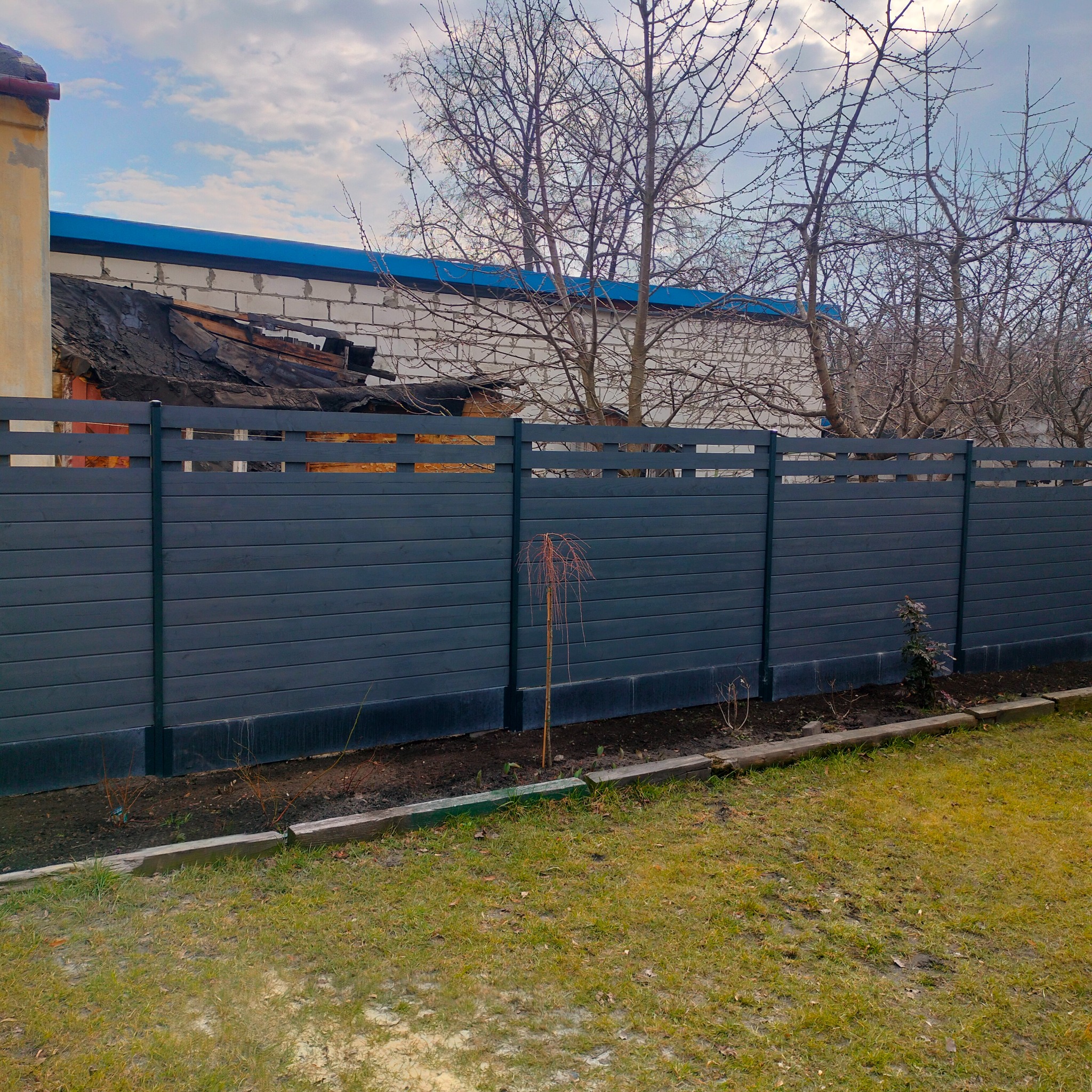 Nowoczesne, grafitowe ogrodzenie panelowe z poziomych desek na tle trawnika, z widoczną konstrukcją domu w tle.