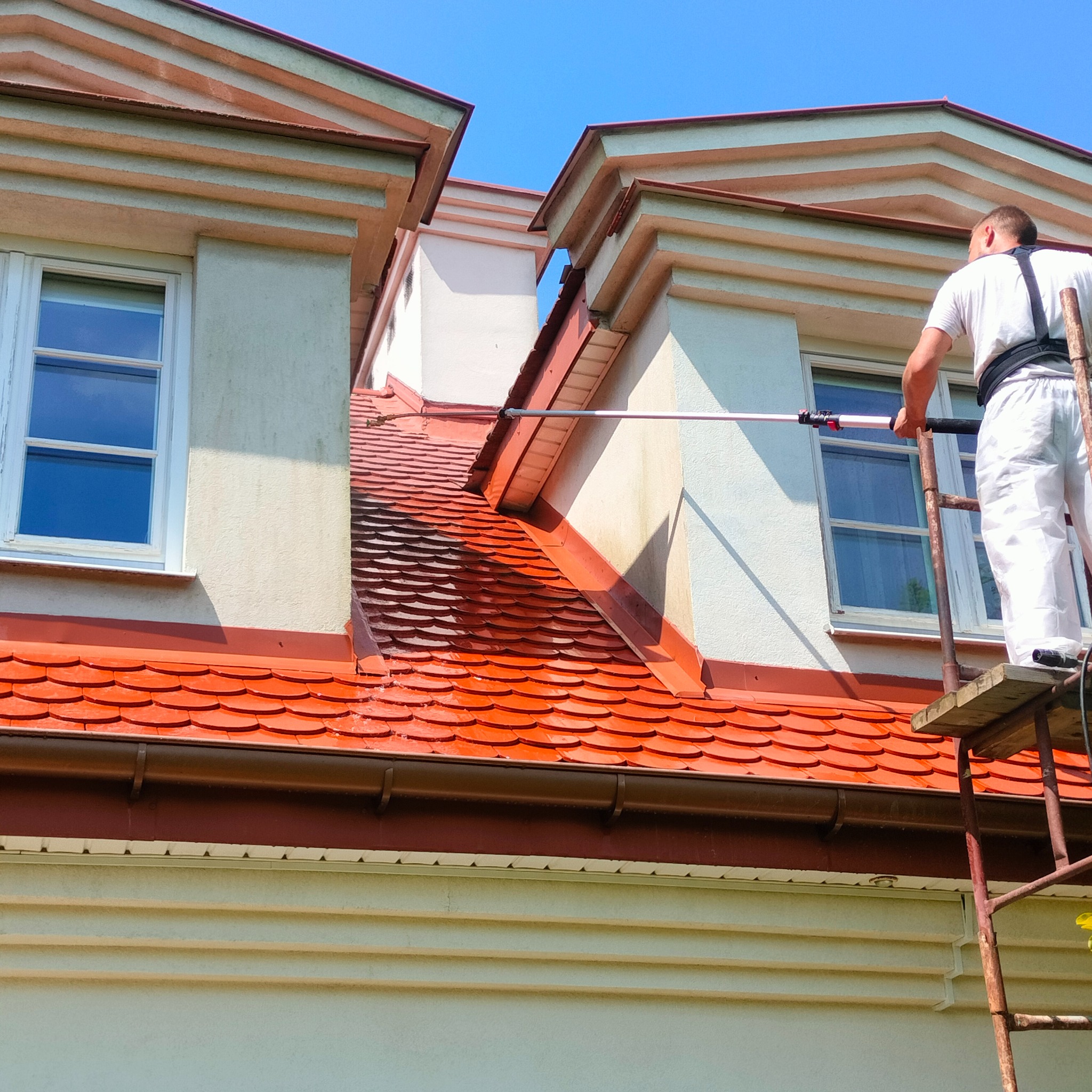 Pracownik w kombinezonie maluje elewację budynku z rusztowania za pomocą długiego wałka. Widoczna pomalowana część dachu i elewacji. Błękitne niebo w tle.