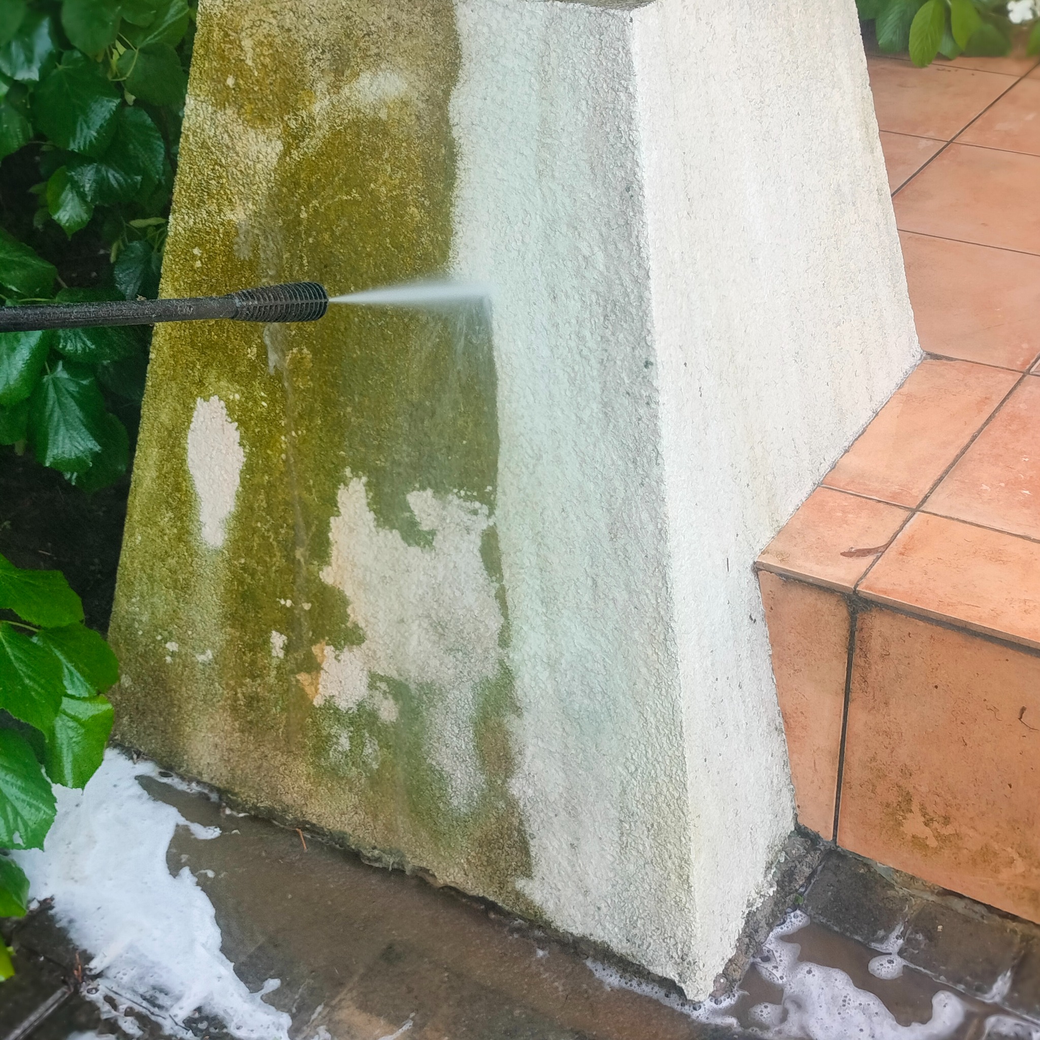 Czyszczenie elewacji myjką ciśnieniową, widoczne efekty przed i po na betonowym filarze przy schodach, zieleń w tle.