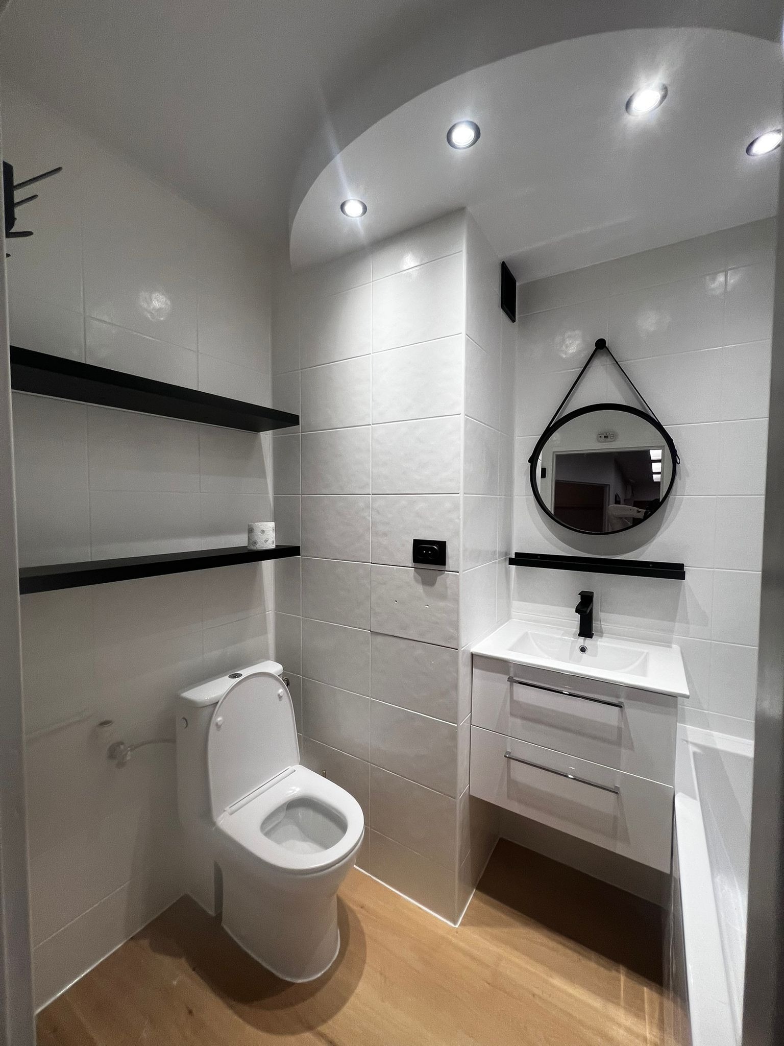 Wykończona łazienka z białymi płytkami, drewnianą podłogą i czarnymi akcentami: półki, okrągłe lustro na pasku i bateria umywalkowa. Oświetlenie punktowe.