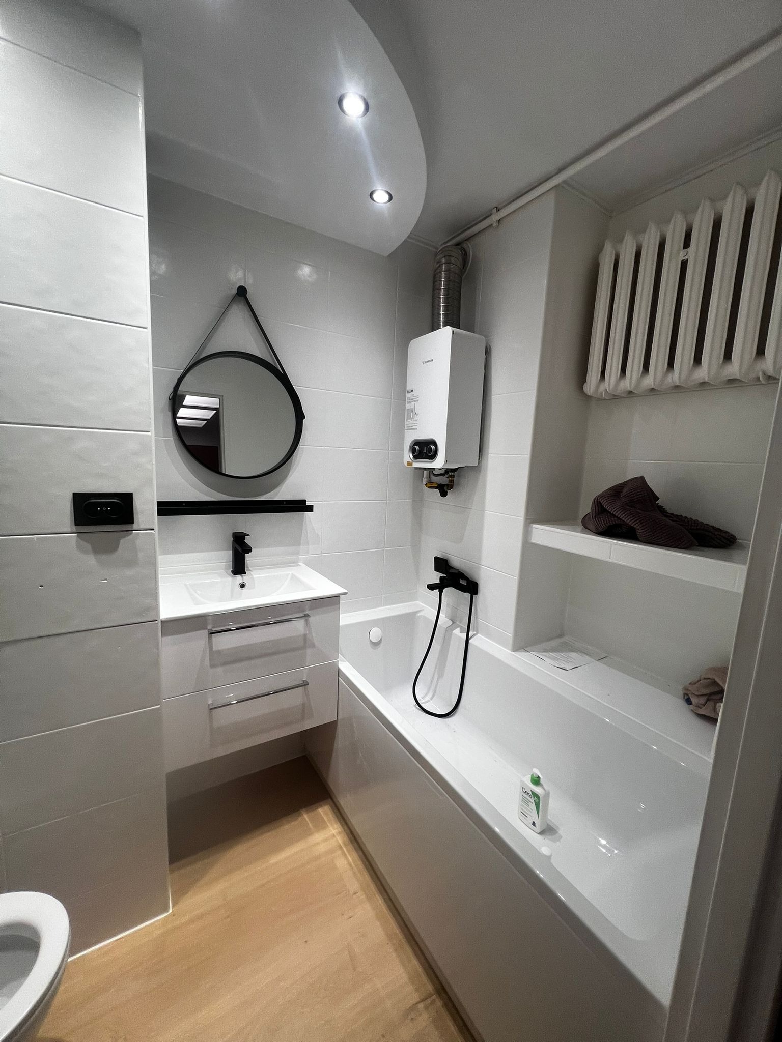 Jasna łazienka po remoncie: białe płytki, wanna z czarną armaturą, okrągłe lustro na pasku i drewniana podłoga. Widoczny bojler i grzejnik.