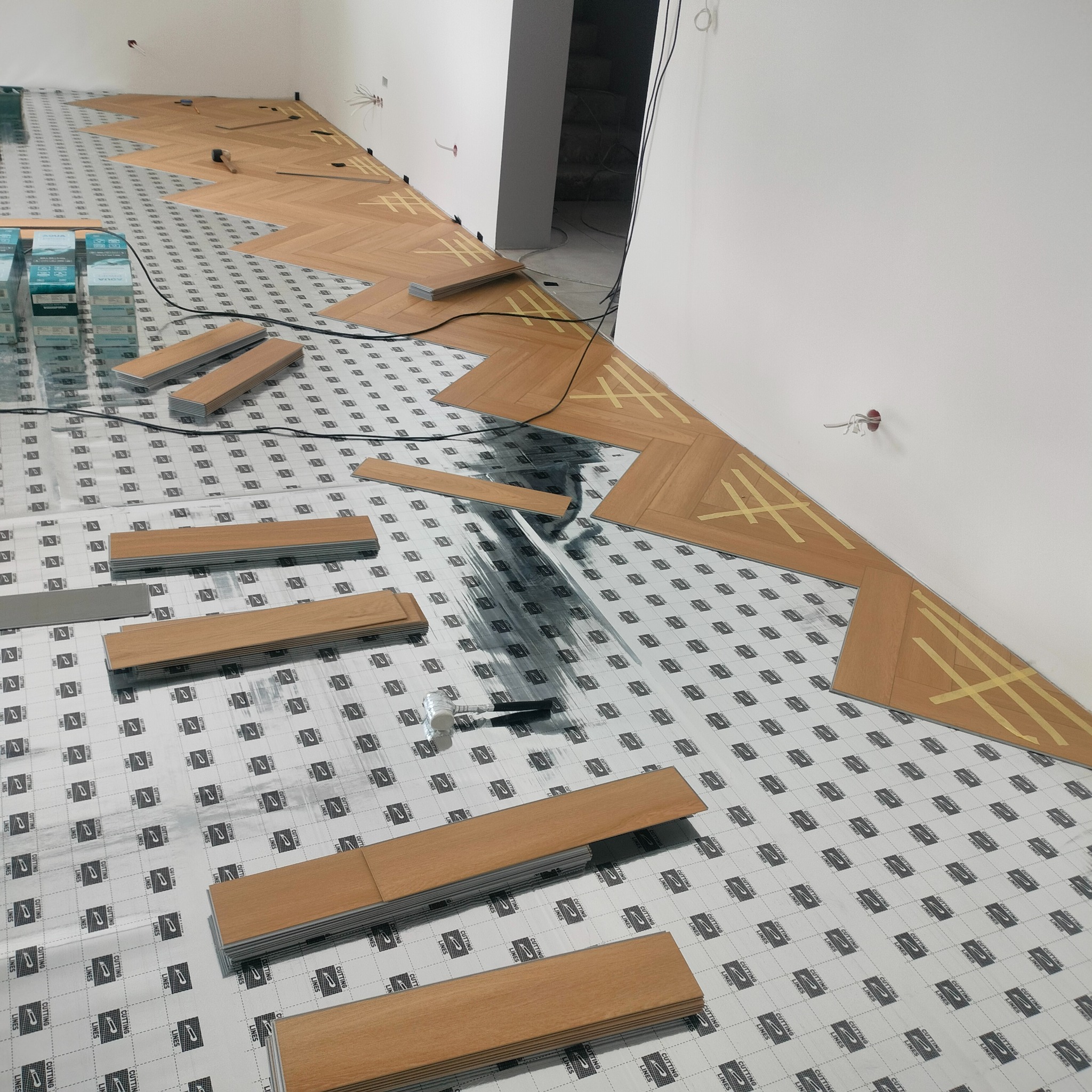 Układanie paneli podłogowych w jodełkę na podkładzie z folii, widoczne narzędzia i pakiety paneli. Częściowo ułożona podłoga w jasnym odcieniu drewna.