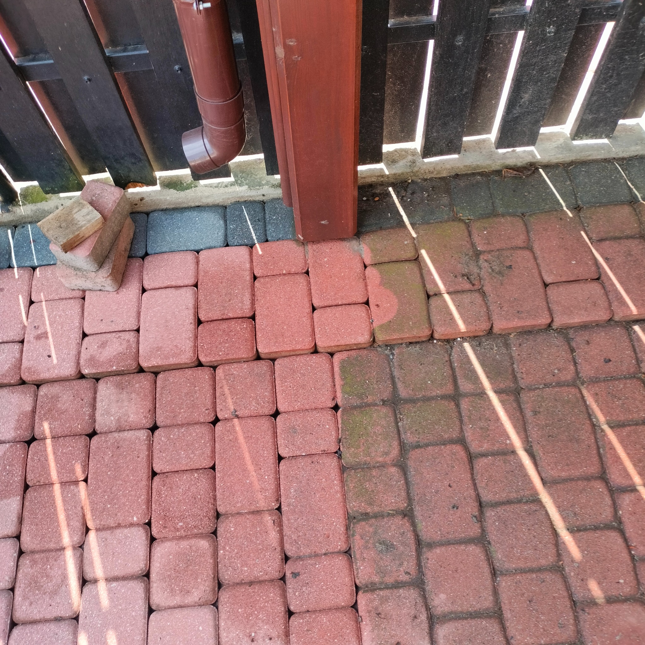 Częściowo ułożona czerwona kostka brukowa przy drewnianym ogrodzeniu i rynnie, widoczne stosy kostki do ułożenia. Fragmenty cienia na kostce.