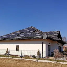 Dom jednorodzinny z panelami słonecznymi na dachu, widoczny zza ogrodzenia w słoneczny dzień.