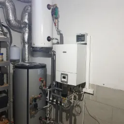 Nowoczesny, biały piec gazowy Bosch z szarym zbiornikiem na wodę Atlantic. Widoczne rury, zawory i przewody. Elementy instalacji grzewczej w kotłowni.